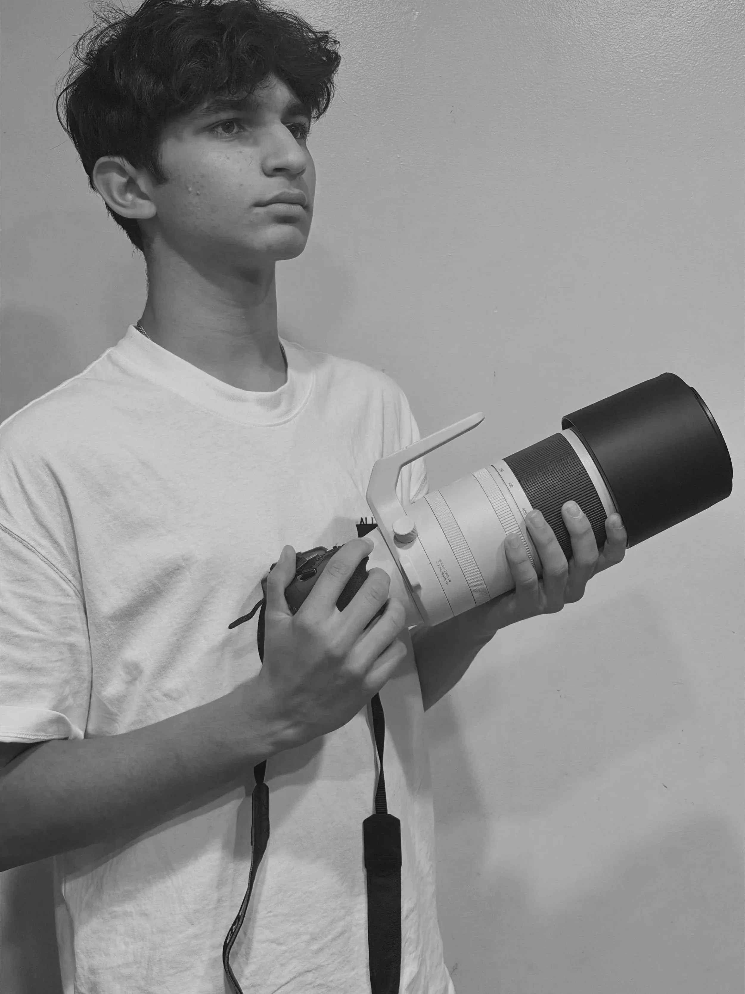 A young person with curly hair wearing a white shirt holds a professional camera with a telephoto lens while standing against a plain wall.