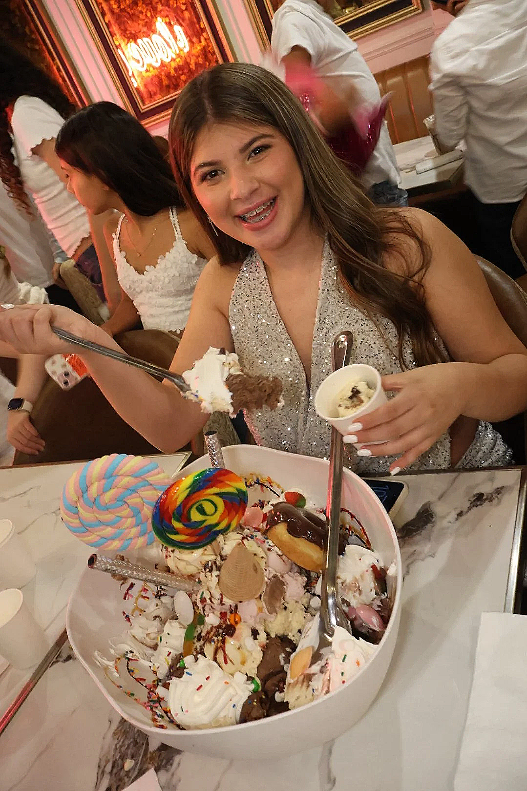 Woman smiling holding a fork and a cup of ice cream at a birthday celebration with a large bowl of assorted ice cream, candies, and toppings in front of her.