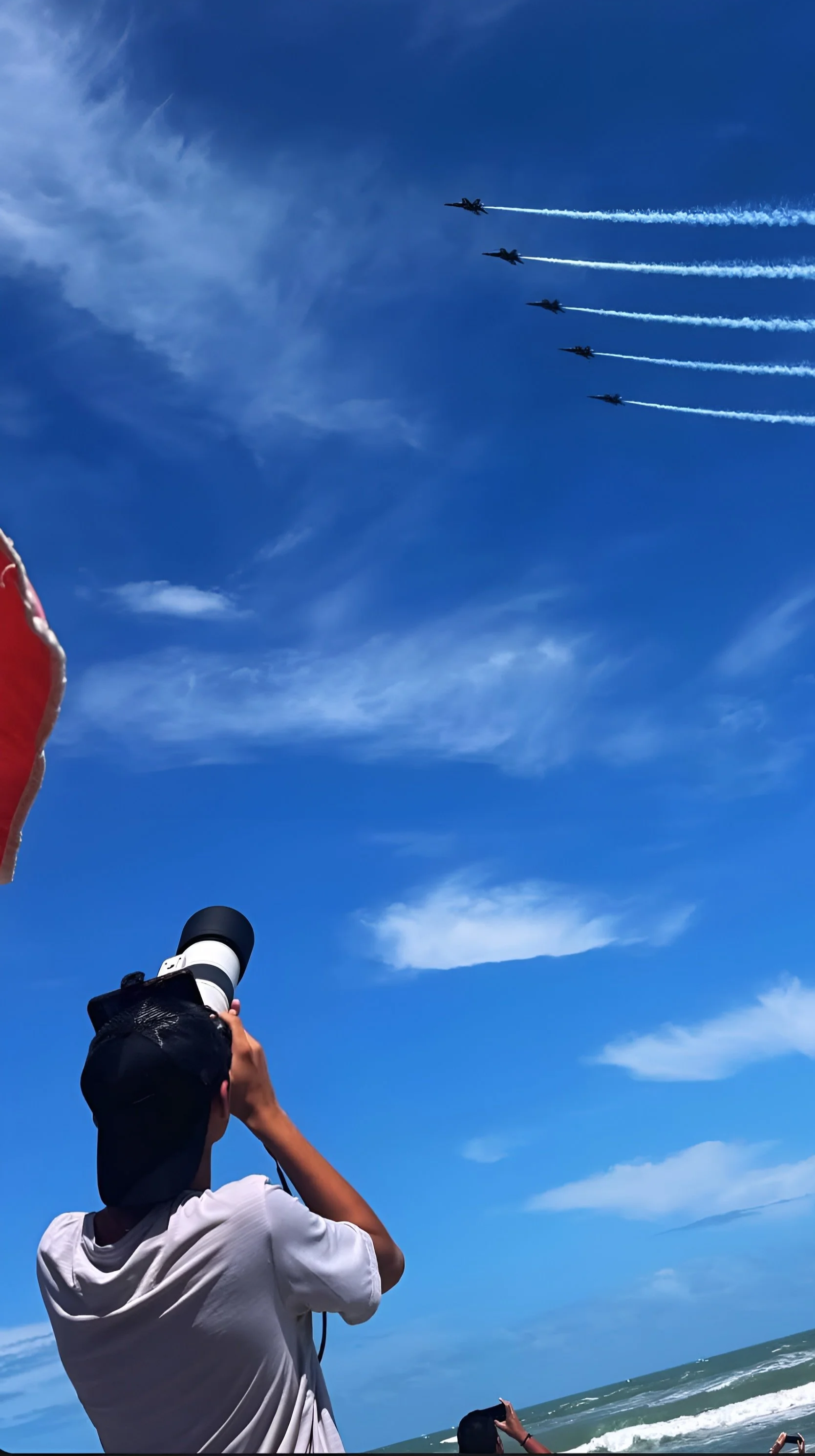 A person at the beach holding a camera and taking photos of a jet performance in the sky, with five jet planes flying in formation leaving white trails behind them on a bright, clear day with a blue sky and some clouds.