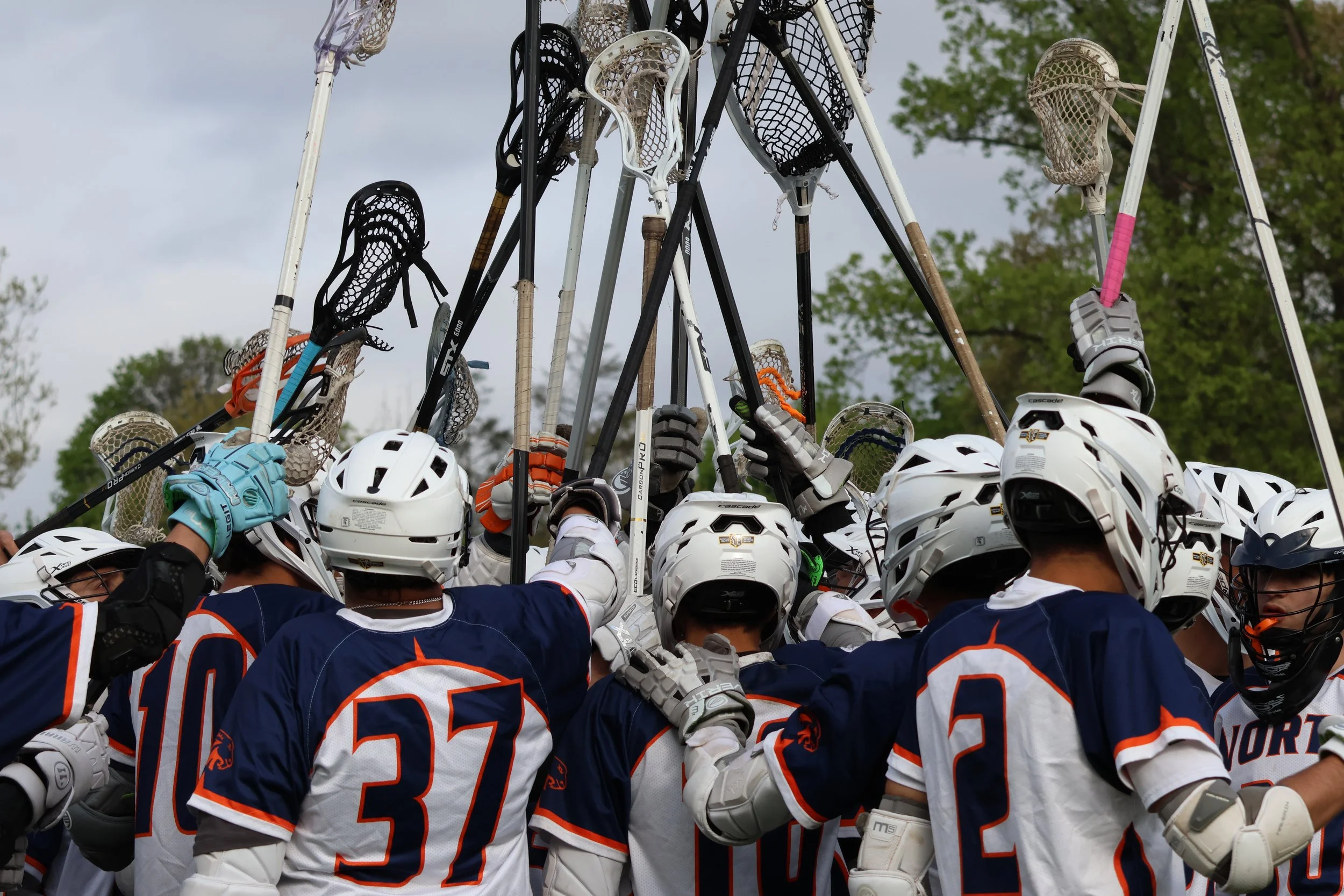 Group of young men's hockey players wearing navy blue jerseys with orange and white accents, white helmets, and gloves, gathered together with their hockey sticks raised above their heads in a celebratory gesture outdoors.