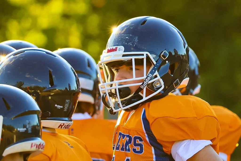 A youth football player wearing a blue helmet and orange jersey with white and blue accents, standing among teammates on a football field during sunset.