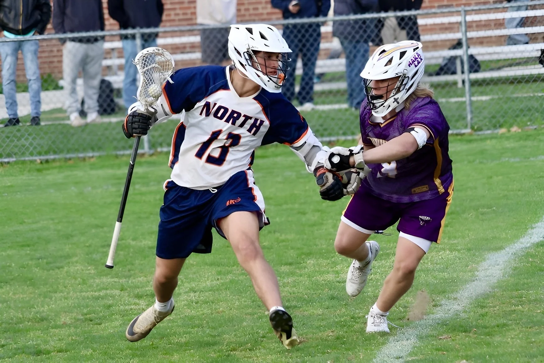 Two girls playing lacrosse outdoors, wearing helmets and sports uniforms, fighting for possession of the ball on a grassy field with a chain-link fence and spectators in the background.