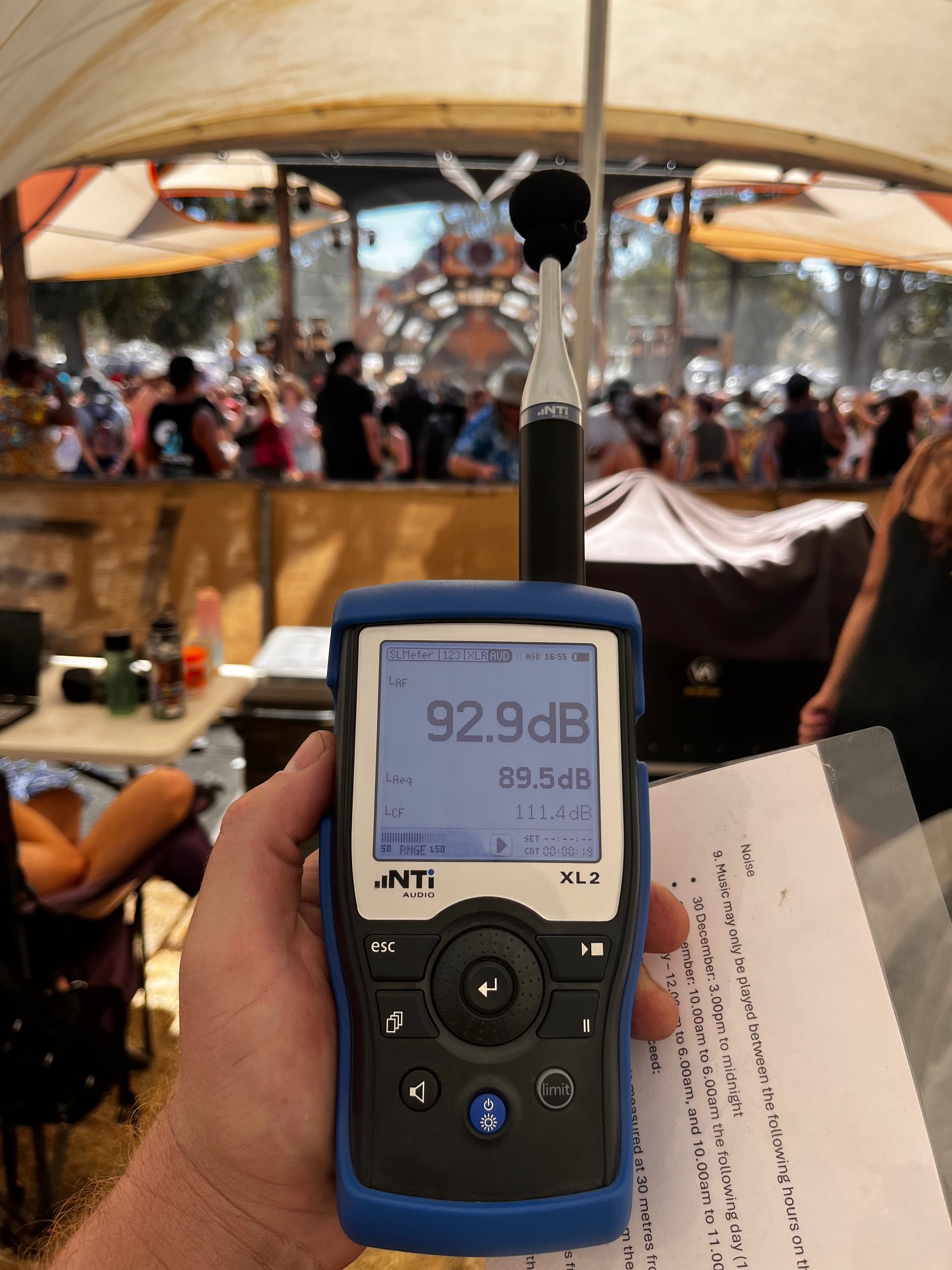 A sound level meter displaying 92.9 decibels, being held by a person's hand with a festival scene in the background showing many people and tents.