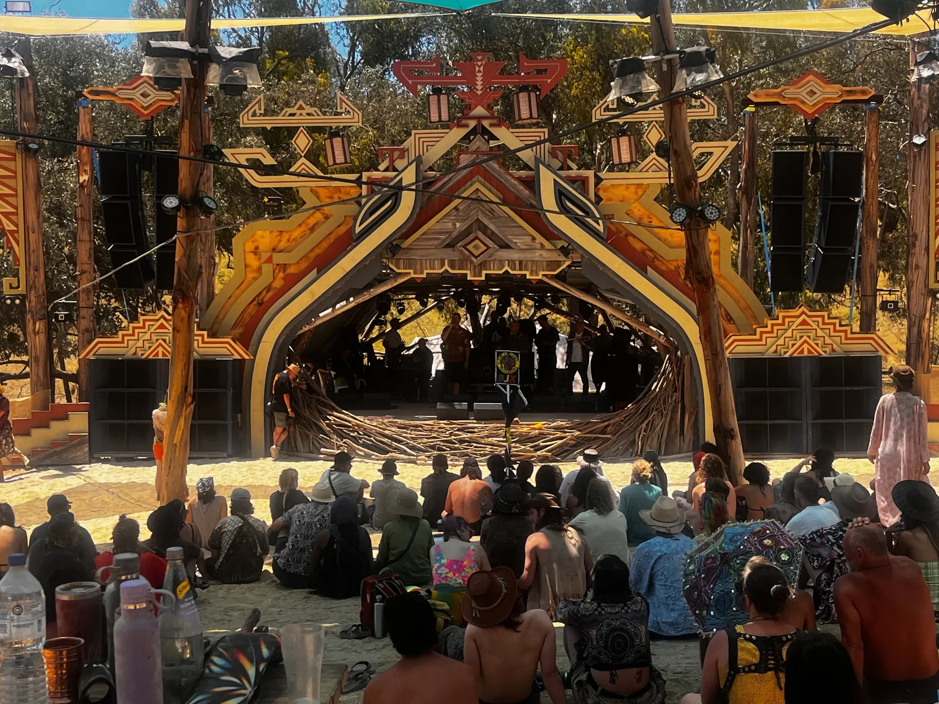 Outdoor stage with native tribal design, wooden poles, and speakers, surrounded by an audience seated on sand, with some standing and taking photos, and bottles of water on a table in foreground.