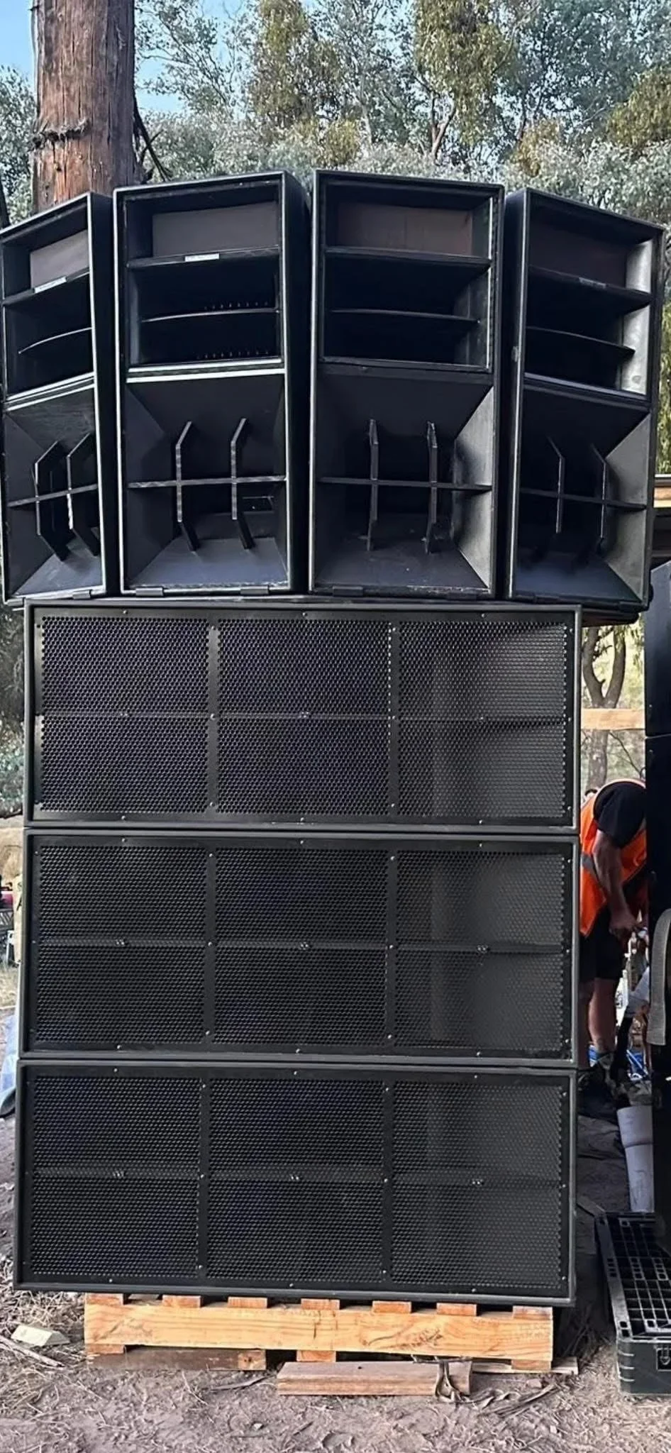 Large black audio speakers stacked outdoors with trees in the background. A person wearing a black shirt and orange safety vest is partially visible on the right side.