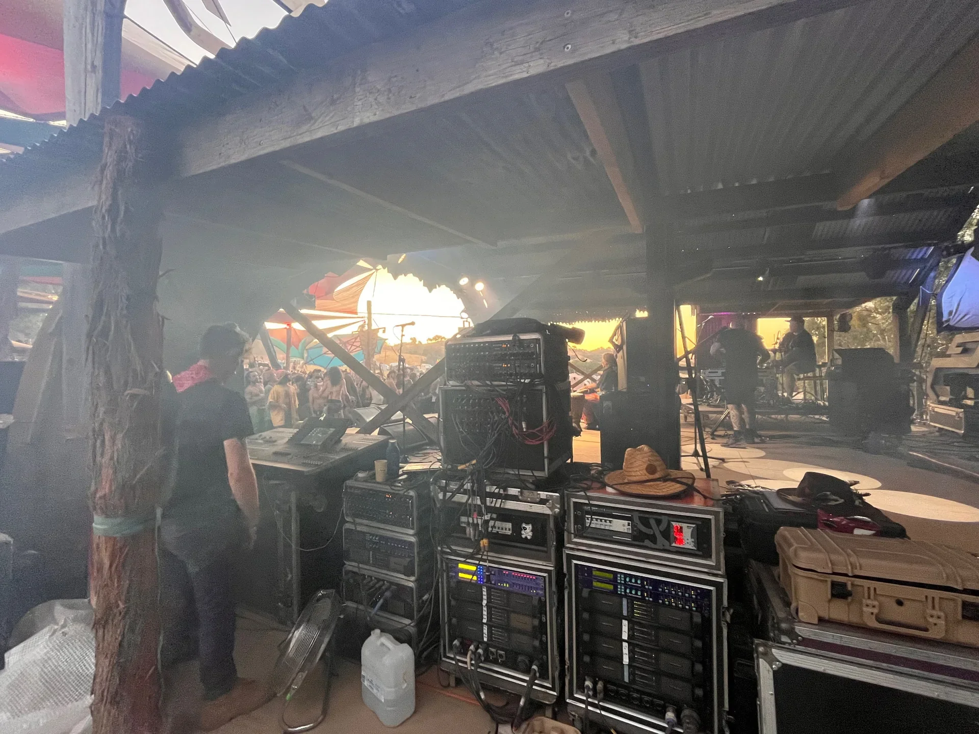 DJ or sound engineer operating sound equipment and mixer at an outdoor event or concert, with a crowd and colorful umbrellas visible in the background.