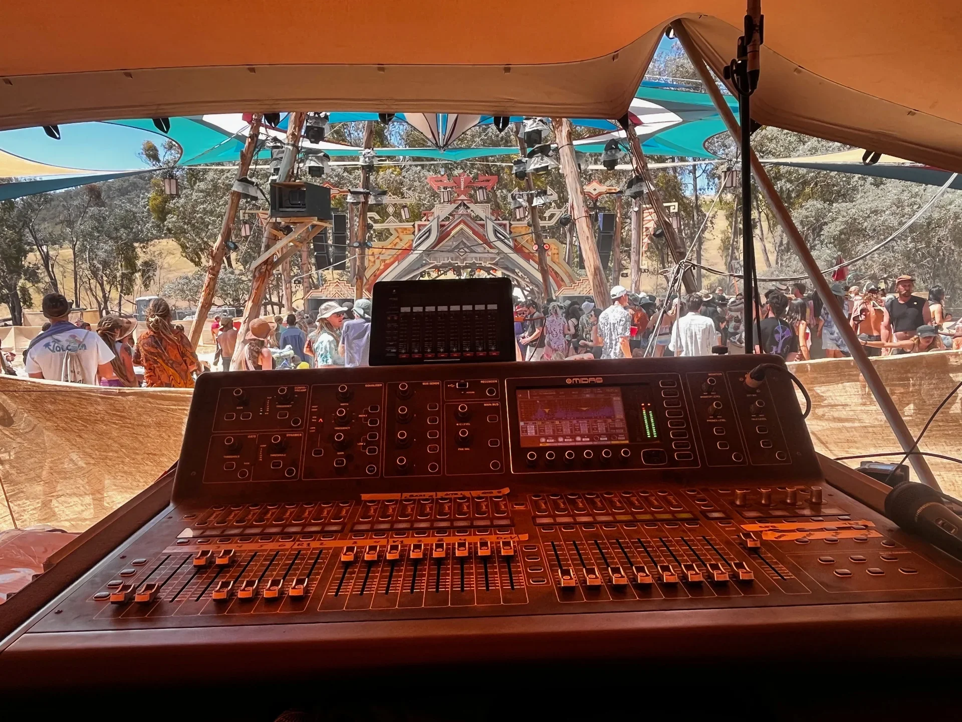 Inside view of a music festival with a DJ booth, DJ equipment, and a crowd of people dancing outdoors under a large canopy with trees in the background.
