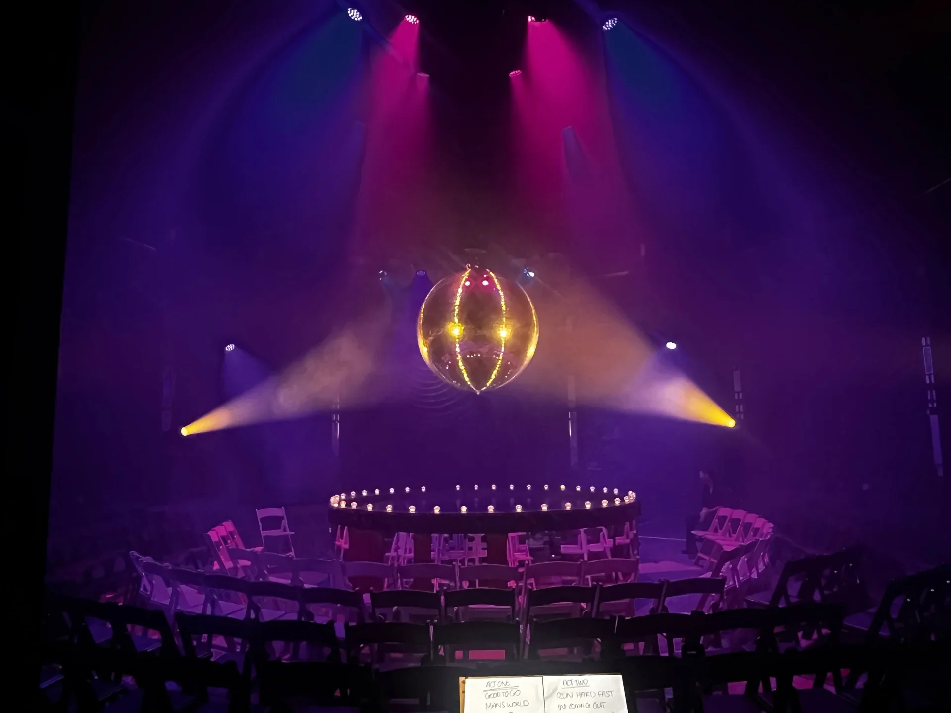 Stage setup with colorful lighting, a disco ball hanging from the ceiling, and chairs arranged in front for an event or performance.