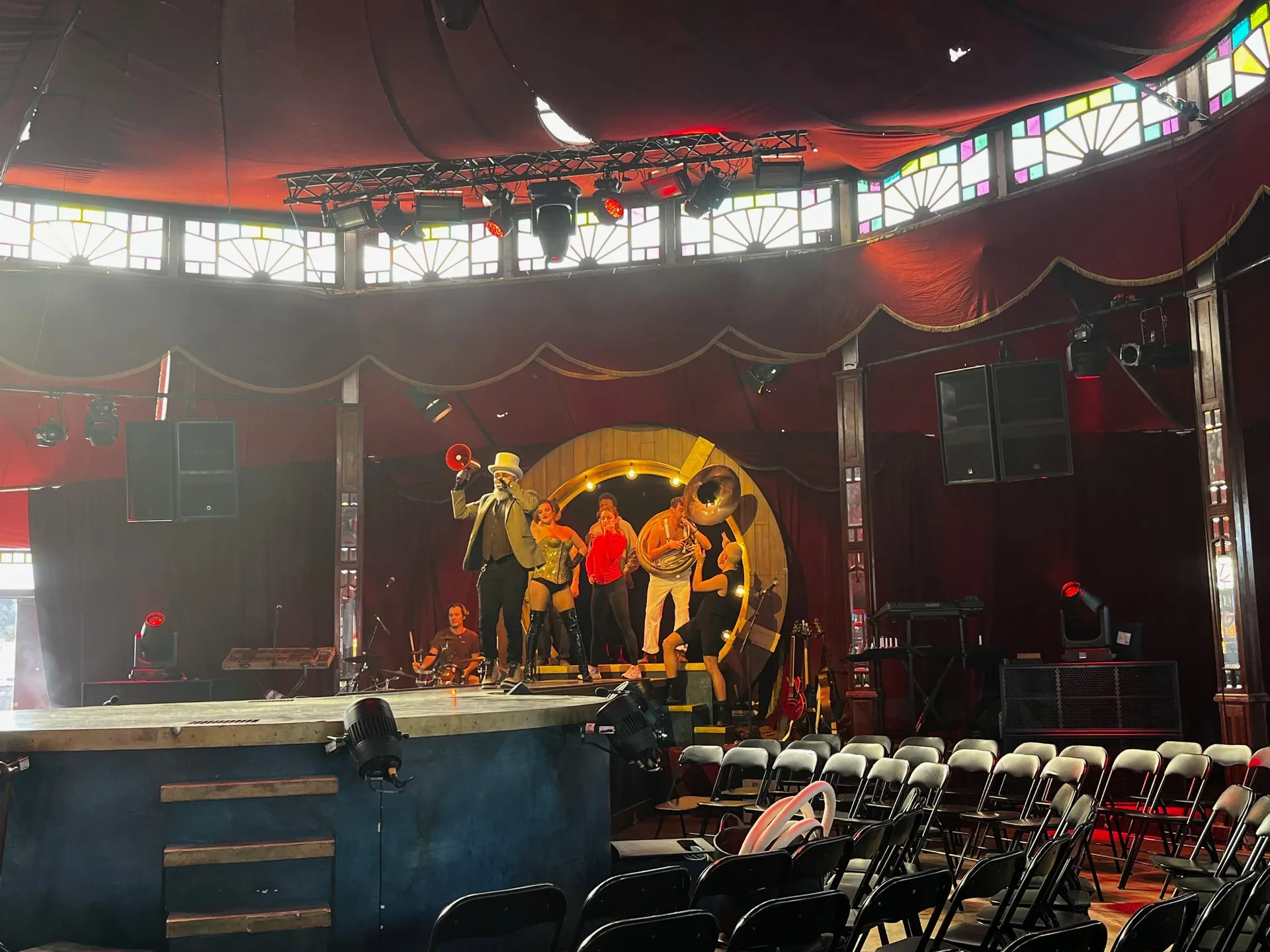 Inside a circus tent with a stage, performers, and security chairs. Bright colored stained-glass windows adorn the upper part of the tent's walls.
