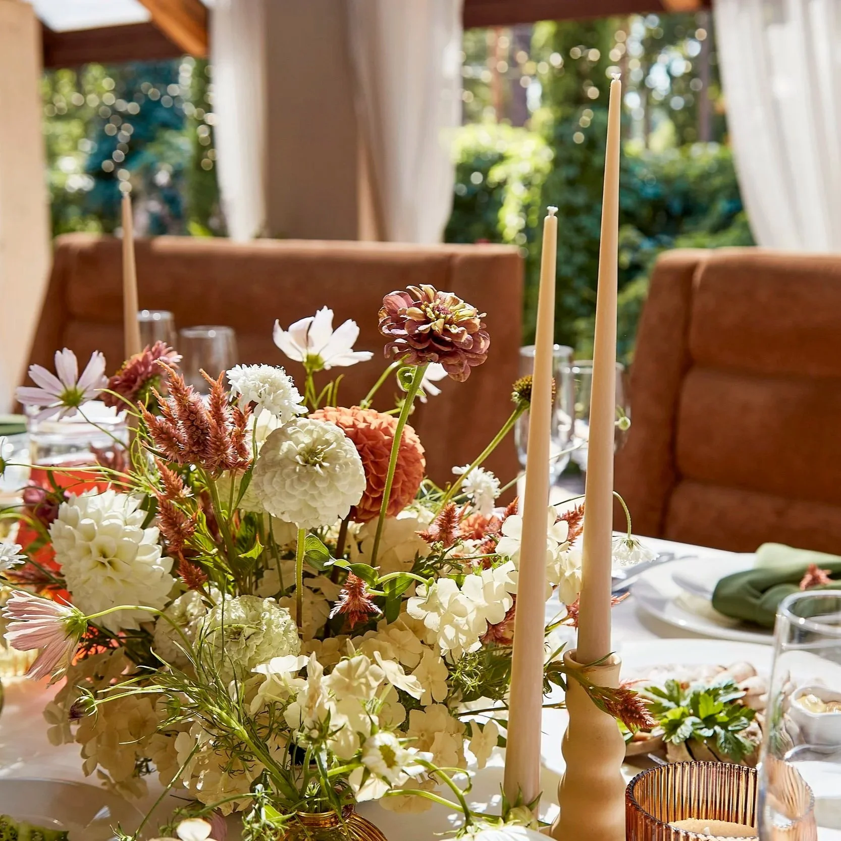 A close-up of a floral centerpiece featuring white, pink, and purple flowers on a dining table set for a celebration. In the background, there is a window with white curtains and a view of greenery outside.
