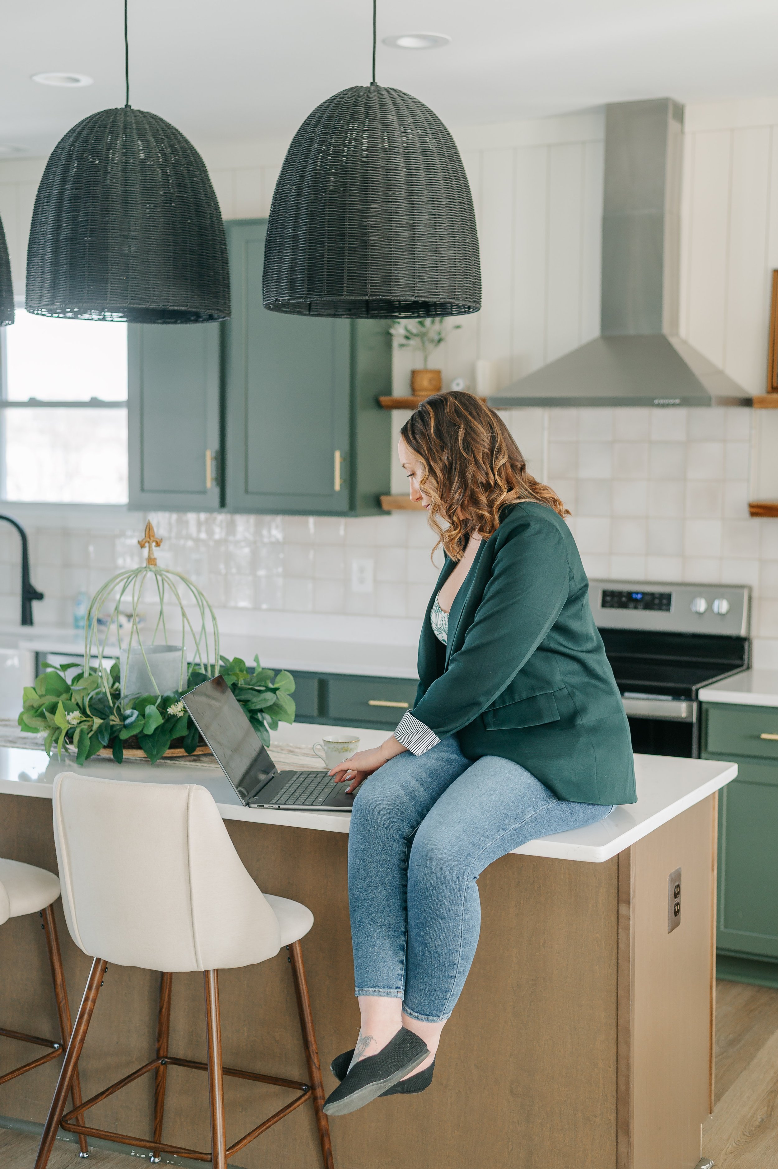 A woman sitting on a kitchen counter working on a laptop, with her right hand on the keyboard. She is wearing a dark green blazer and blue jeans. The kitchen has green cabinets, a white tiled backsplash, and two large black woven pendant lights hanging from the ceiling. There is a plant arrangement in the foreground and a ceiling vent is visible.