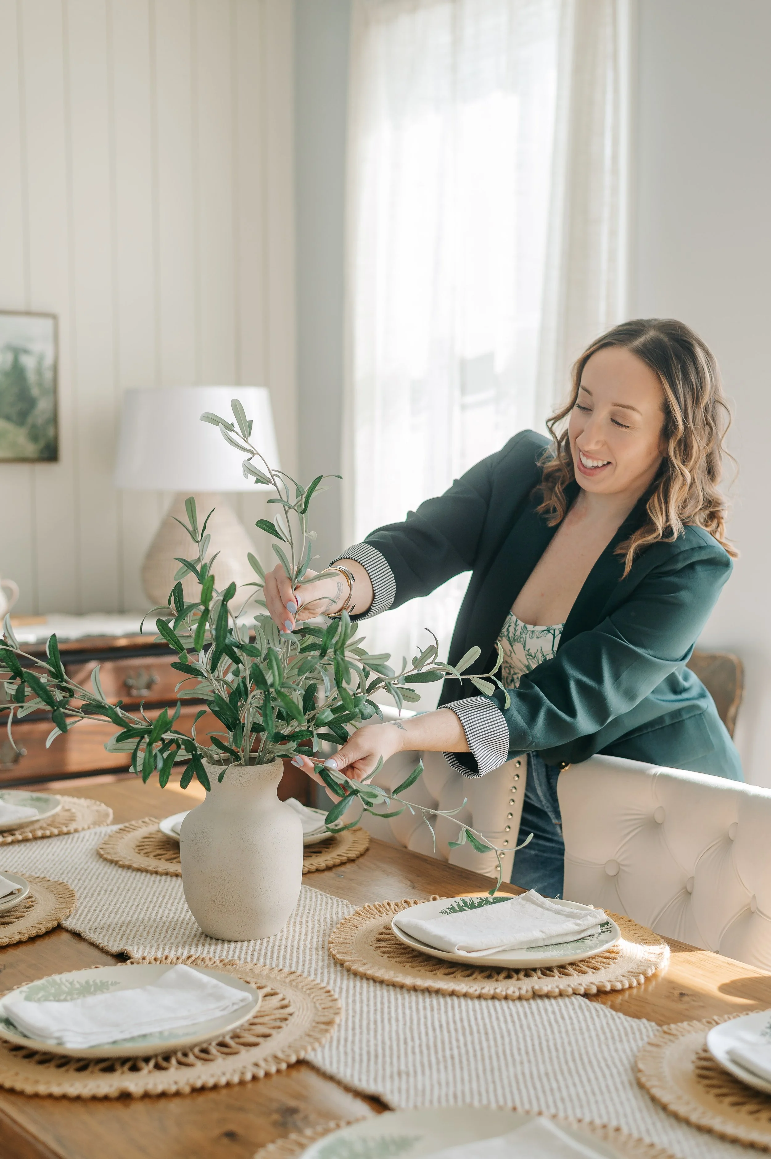 A woman arranging a potted olive tree on a dining table with place settings, in a bright, cozy dining room with natural light.