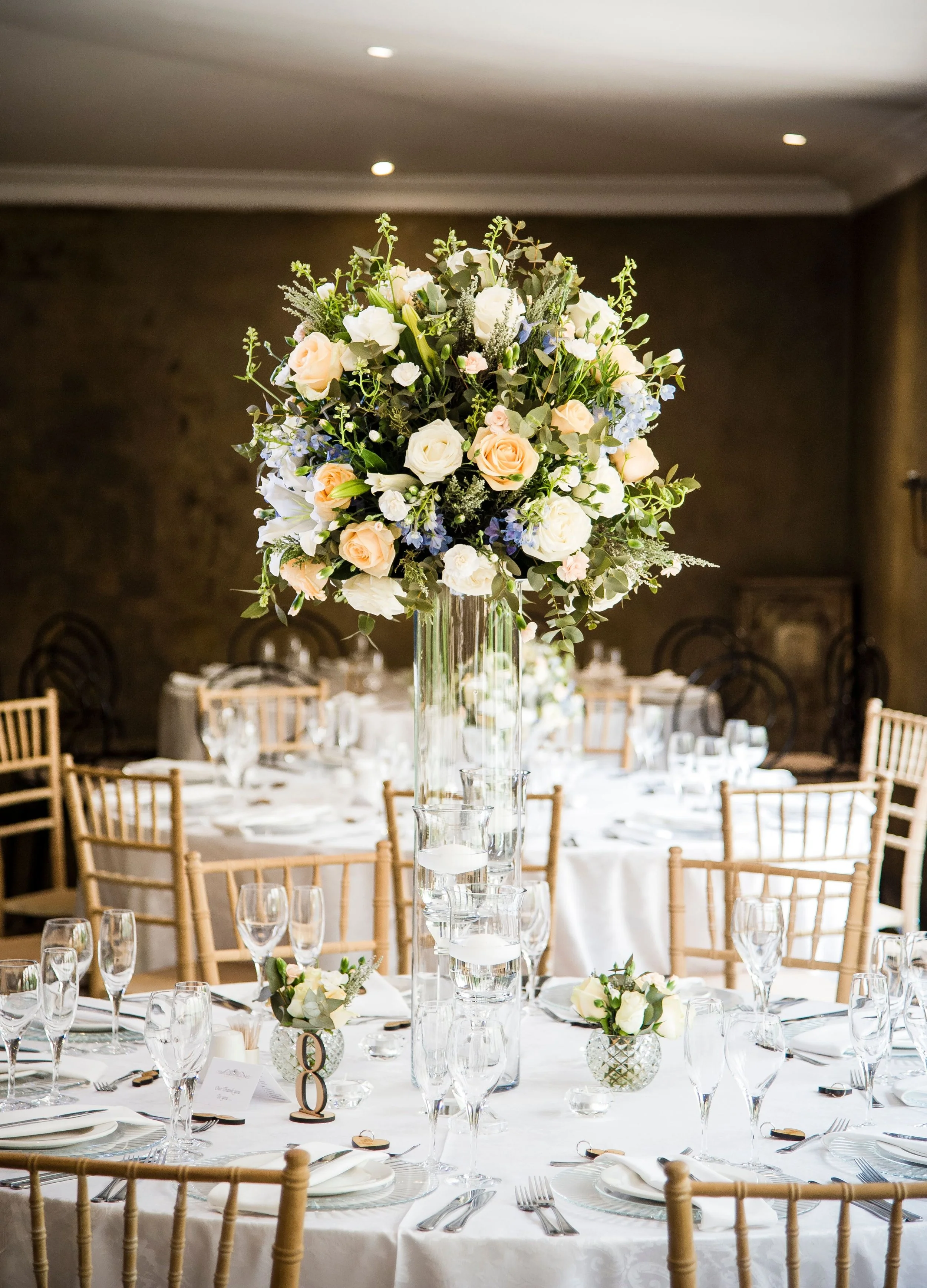 A formal event table decorated with a tall floral centerpiece consisting of white and peach roses, white lilies, and greenery in a glass vase. The table is set with multiple wine glasses, plates, utensils, and small floral arrangements in glass containers. The background shows a dark wall and chairs arranged around the table.