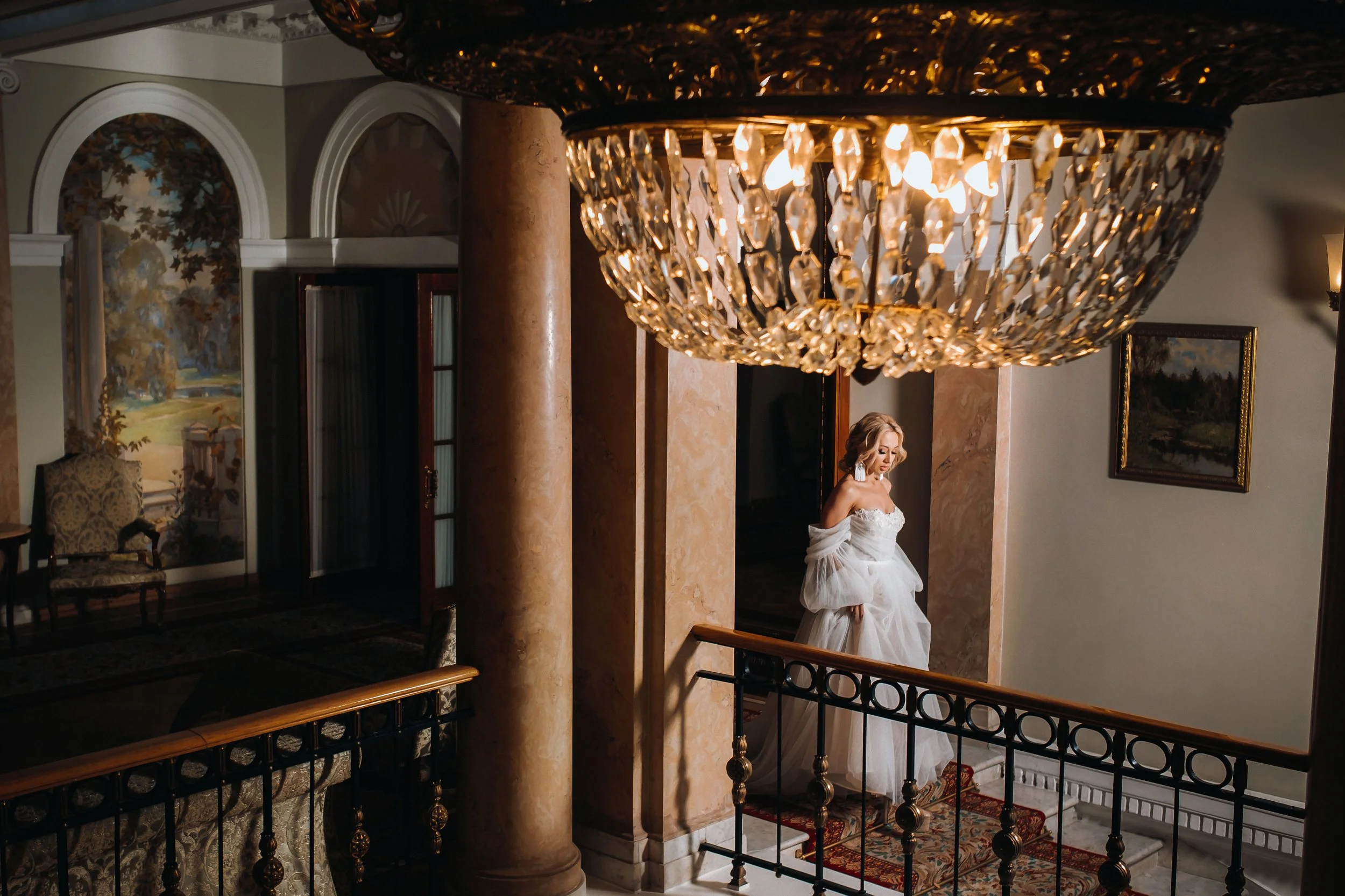 A woman in a strapless white wedding dress with a ruffled skirt standing on a staircase in an ornate interior, illuminated by a large chandelier above.