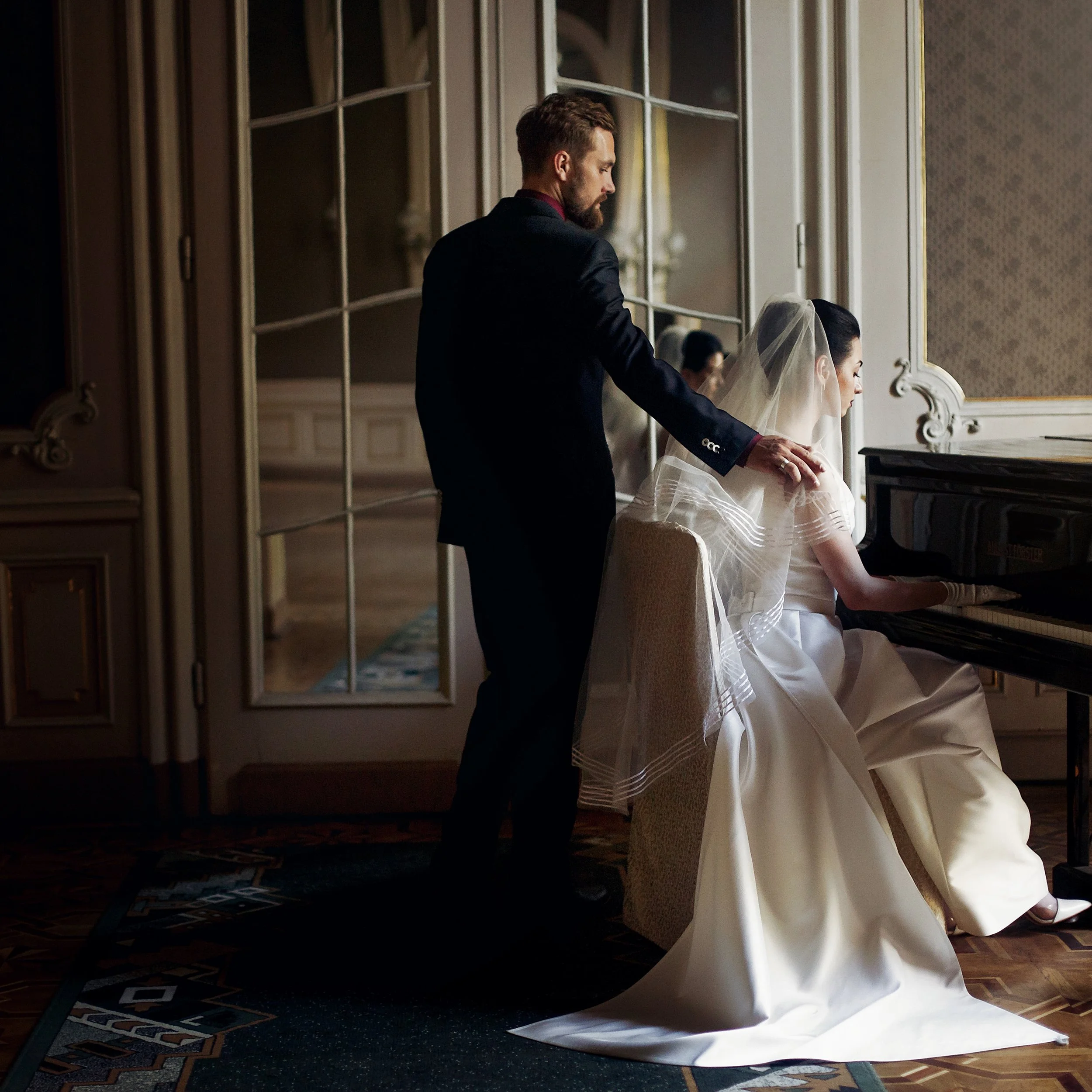 A groom in a dark suit stands beside a seated bride in a flowing white wedding gown with a veil, holding her hand during a wedding ceremony in an elegant room with large windows and ornate décor.