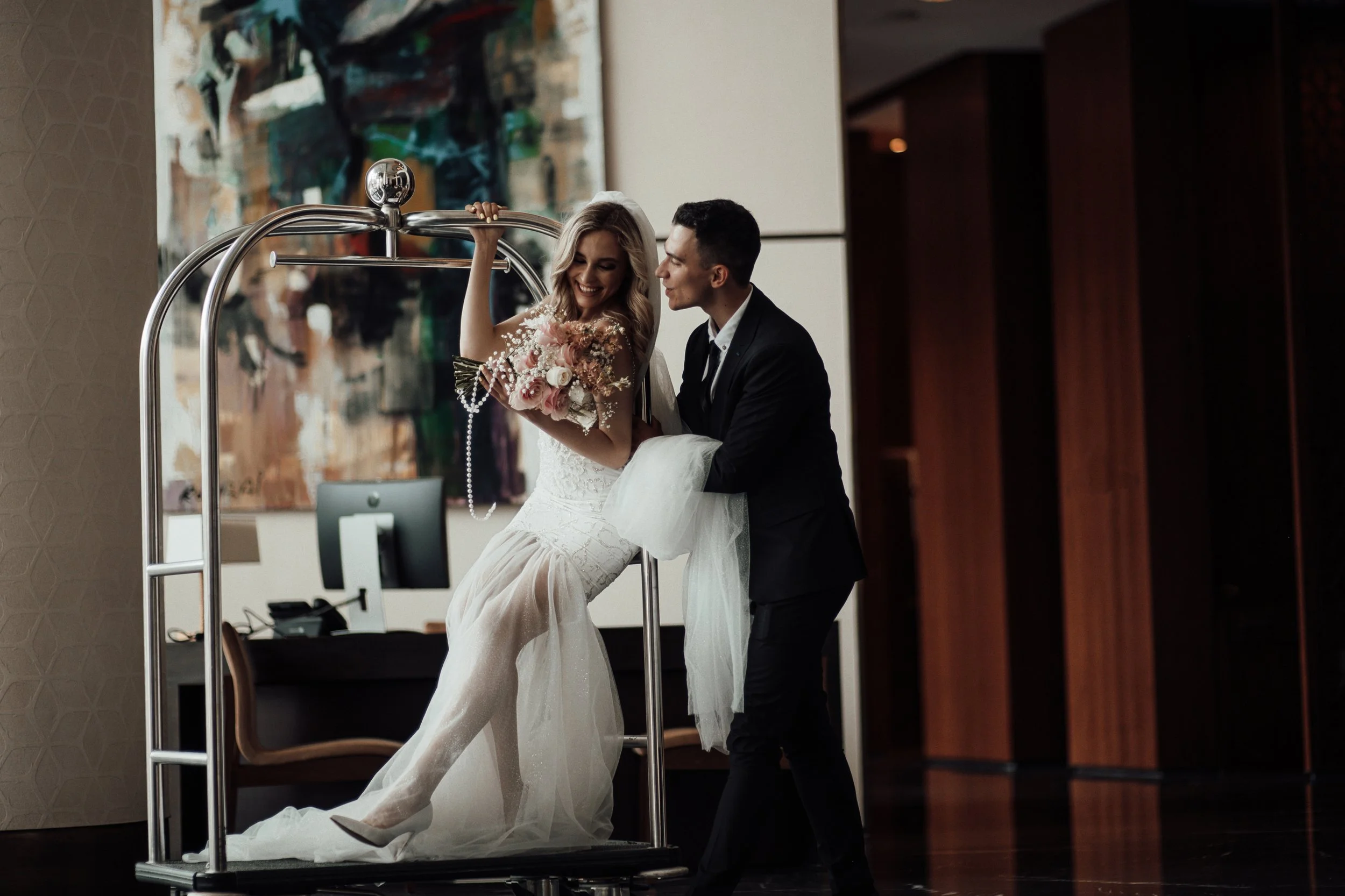 A bride in a white wedding dress holding a bouquet of pink and white flowers, smiling and leaning on a luggage cart, while a groom in a dark suit stands beside her. They are indoors, next to a large painting and a wood-paneled wall.
