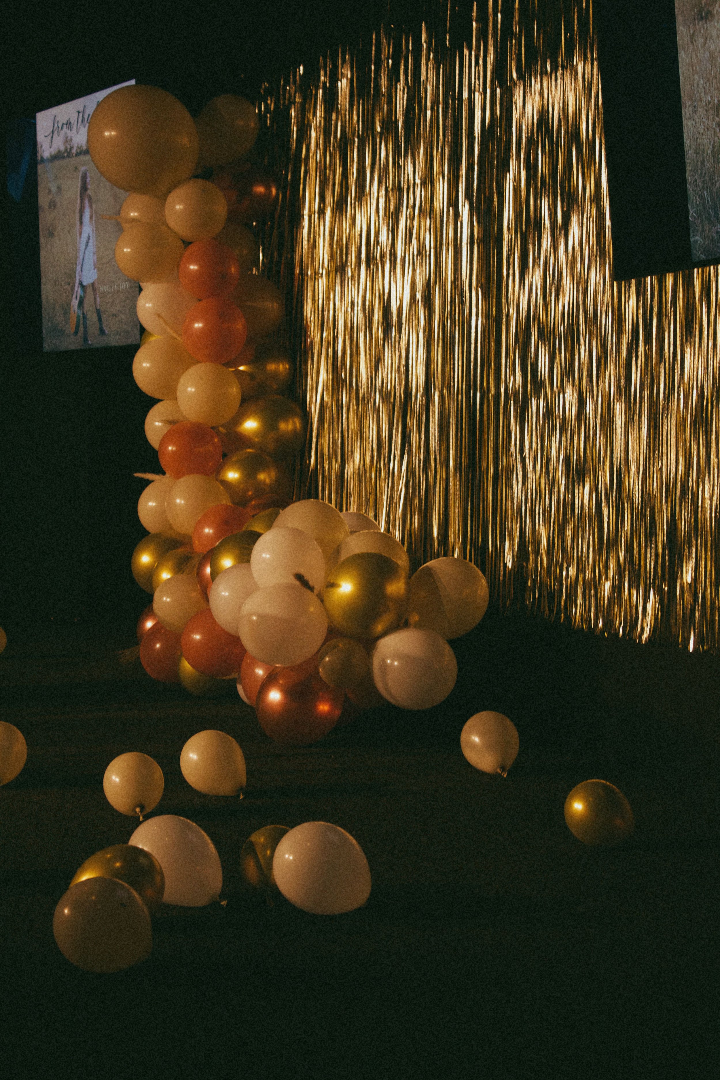 A decorative scene with gold and white balloons, a gold curtain backdrop, and a small sign partially visible in the background.