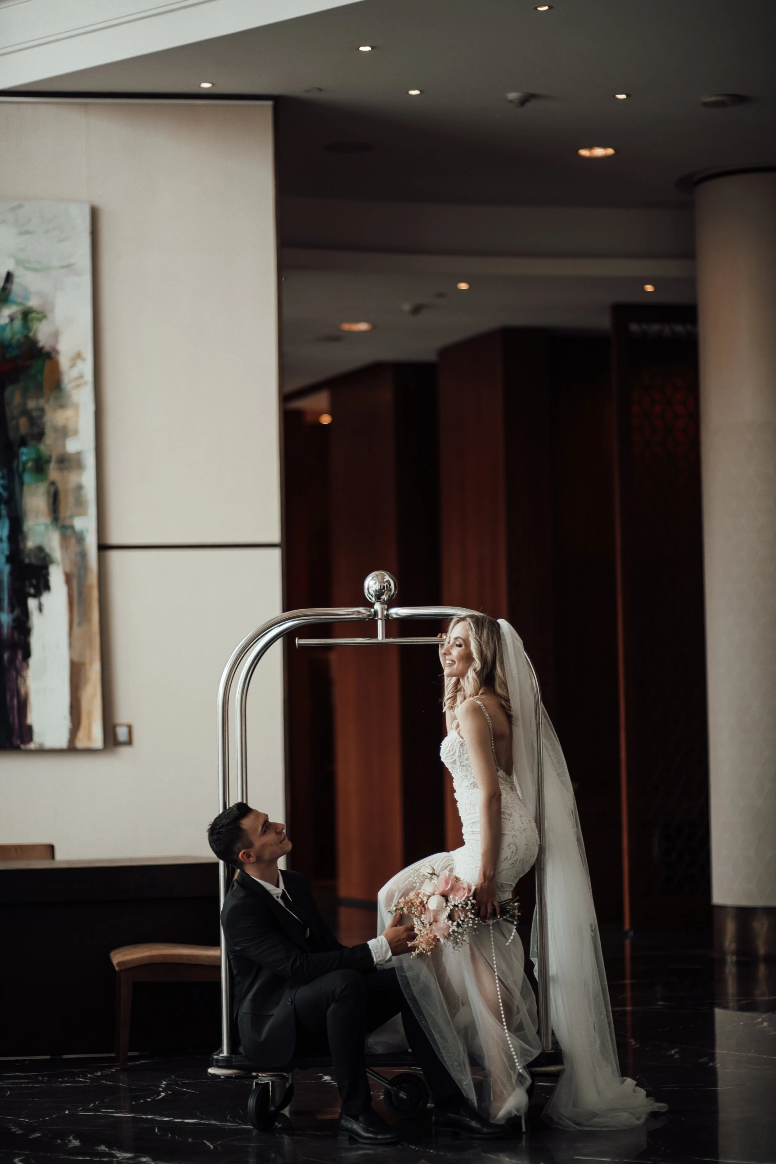 A bride with long blonde hair in a white wedding dress and veil sits on a medical gurney, holding a bouquet of pink and white flowers. A groom in a black tuxedo is kneeling in front of her, smiling and holding her hand in a hospital or hotel lobby with dark marble floors and art on the wall.