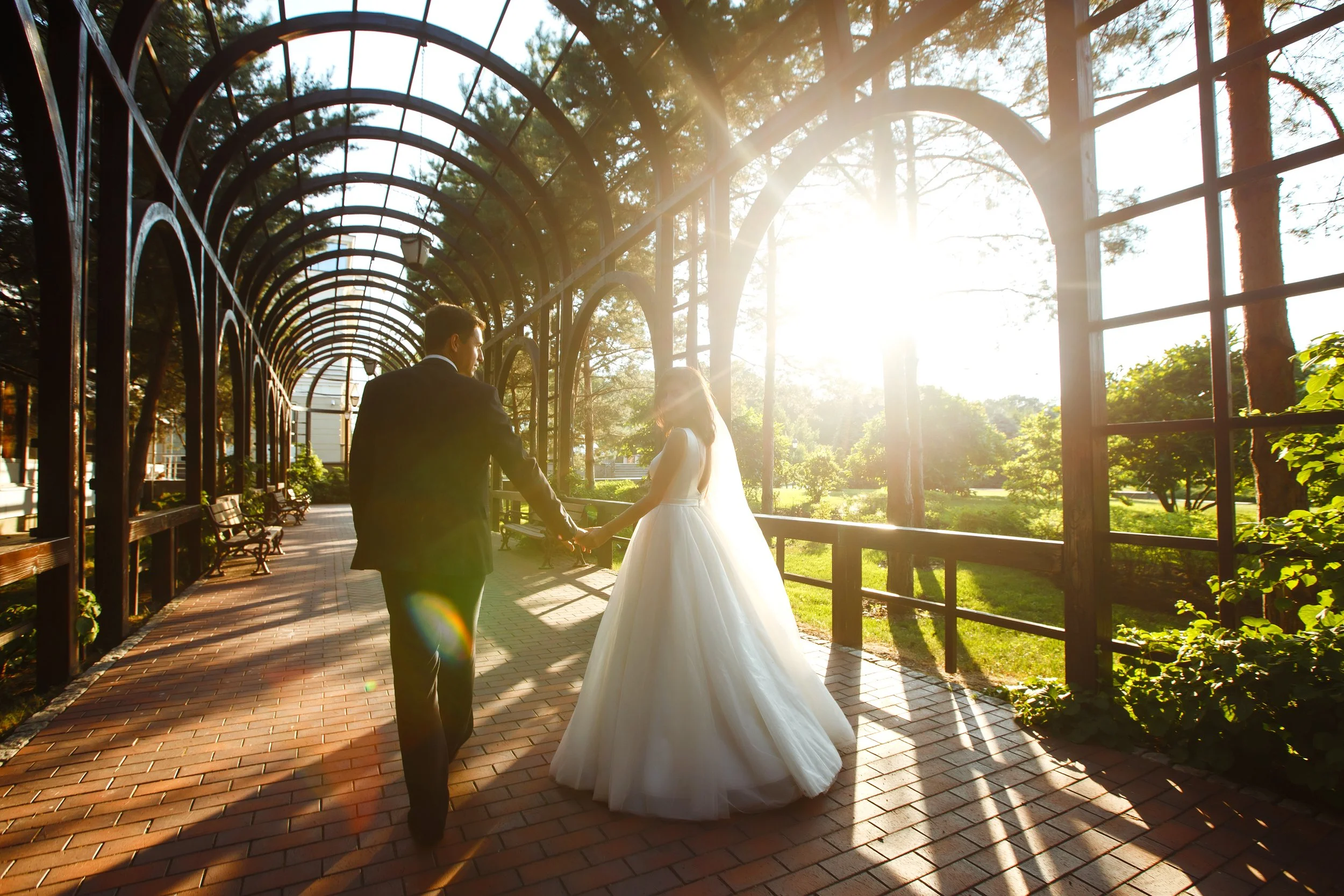A bride and groom holding hands and walking under a metal archway with sunlight shining through. The scene is outdoors in a park or garden setting with trees and greenery.