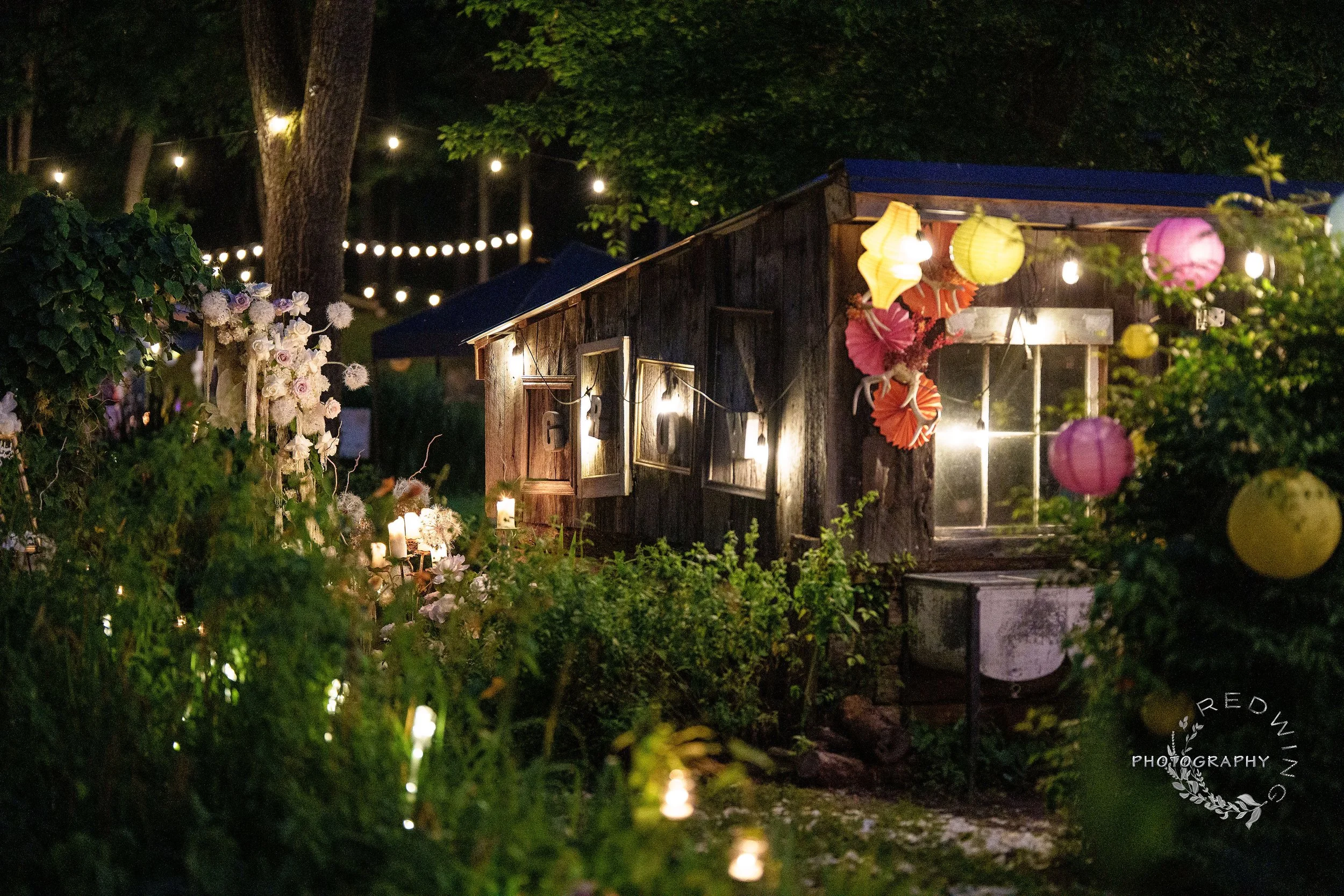 Night scene of a rustic wooden shed decorated with colorful paper lanterns and string lights, surrounded by flowers and greenery.