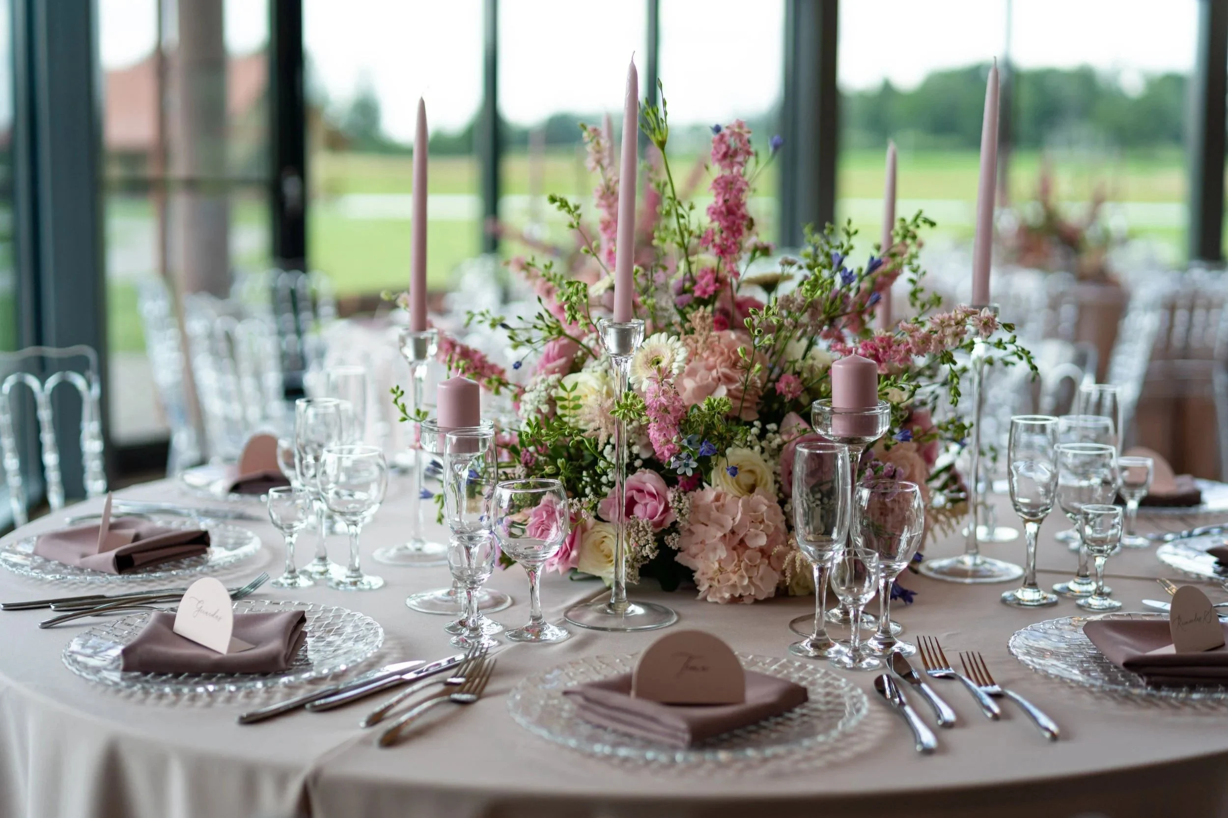 A round banquet table decorated with a large pink and white floral centerpiece, surrounded by glasses, silverware, and folded mauve napkins with place cards in a bright, open room with large windows and a view of greenery outside.