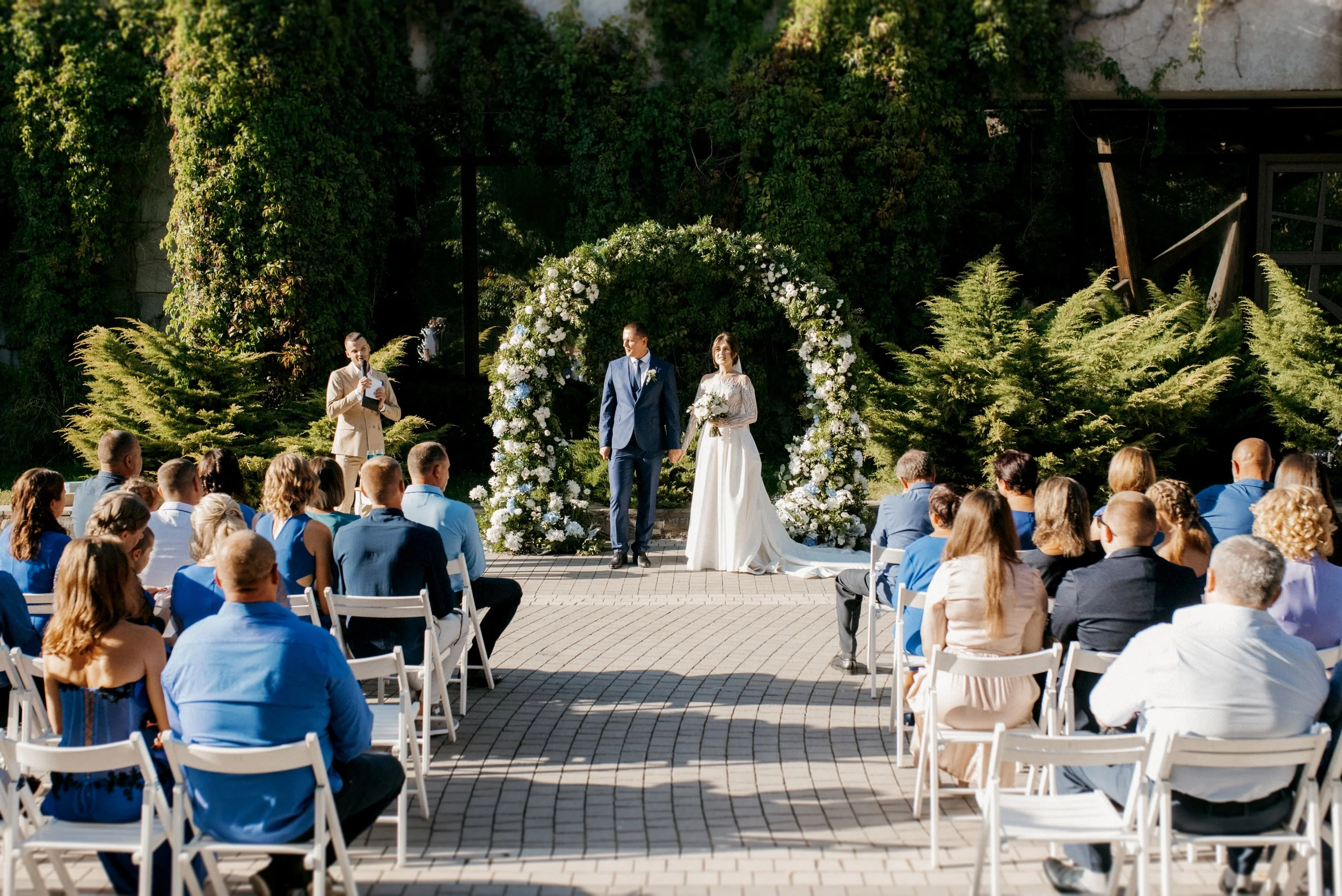 A wedding ceremony outdoors with a couple standing under a floral arch, surrounded by guests seated on white chairs, greenery, and trees.