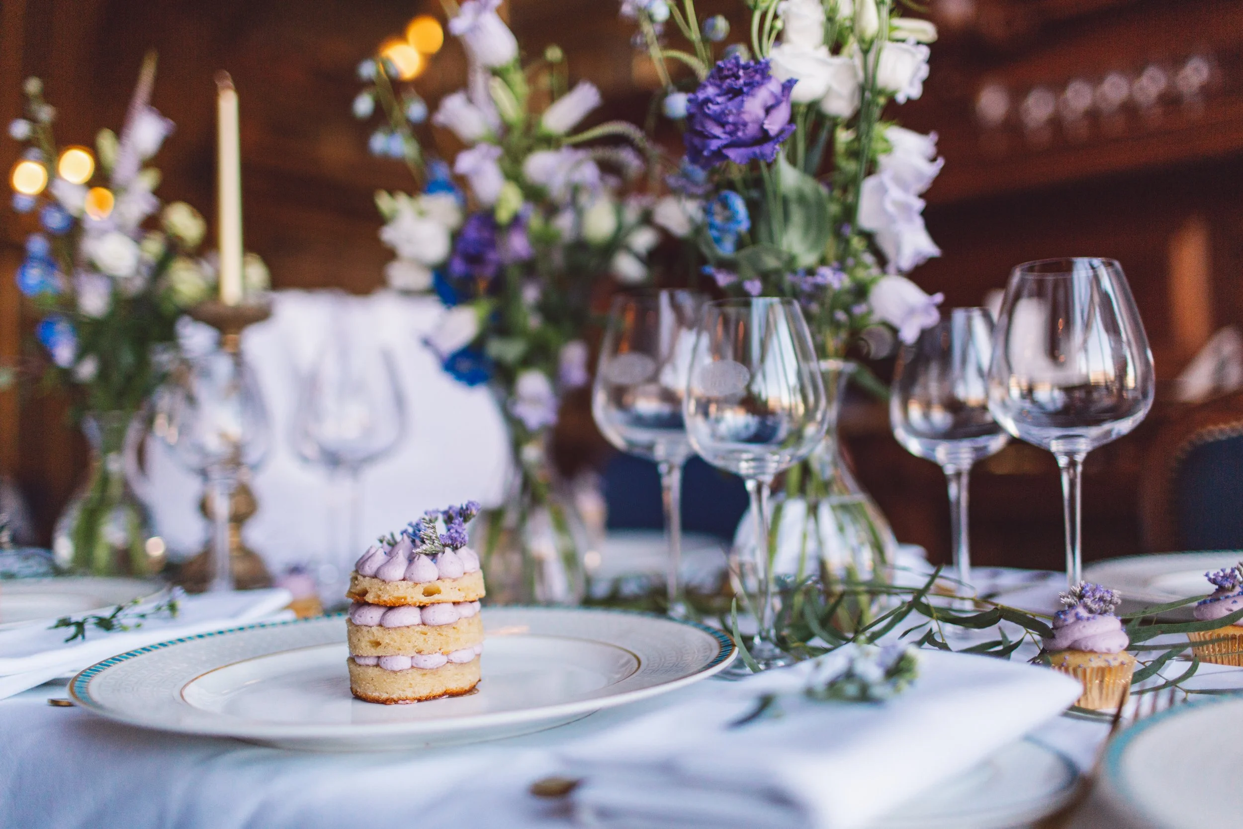 A table setting with a floral centerpiece featuring purple and white flowers, elegant glassware, and a small layered dessert with purple cream on a decorative plate.