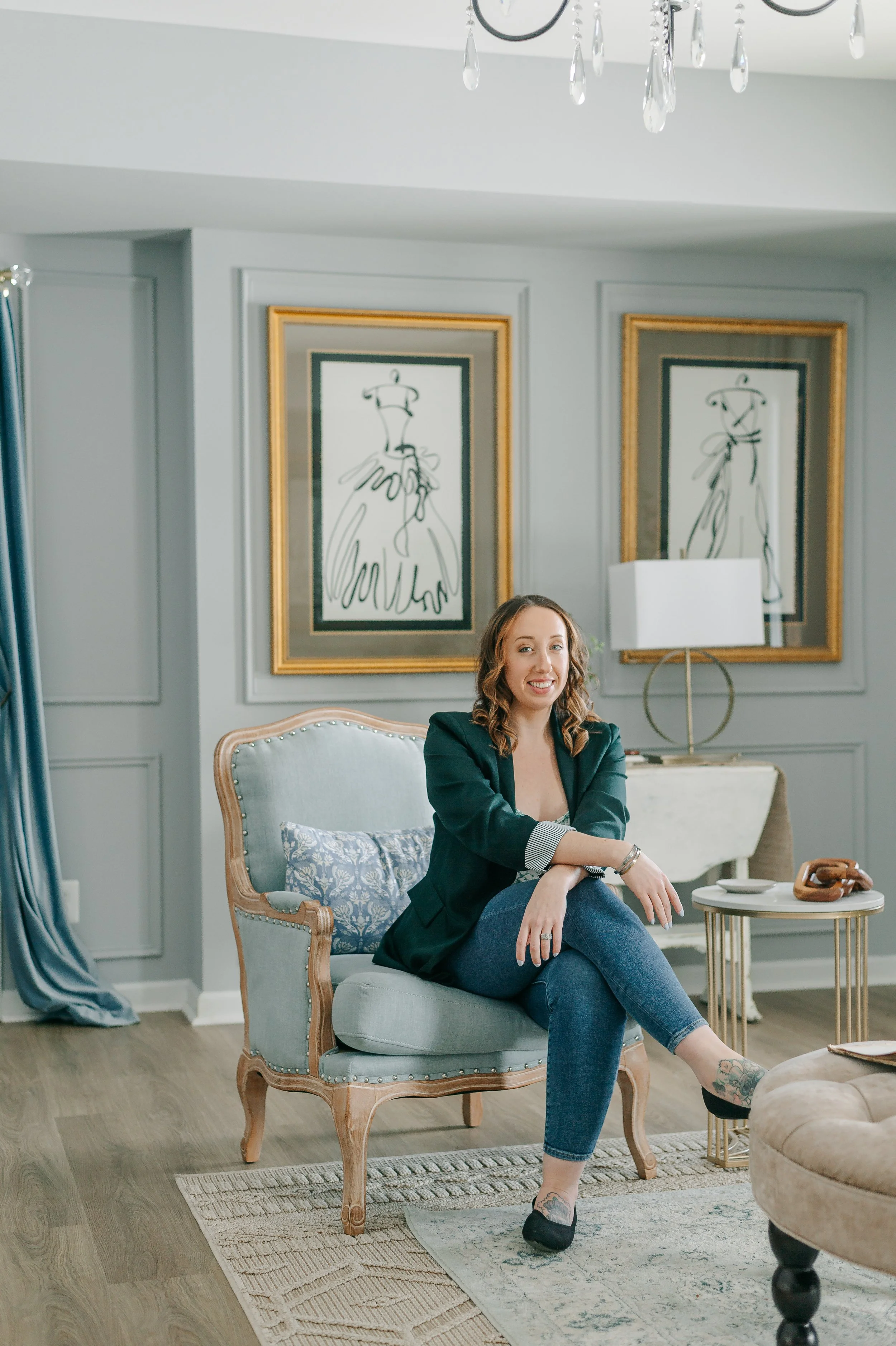 A woman with wavy brown hair and tattoos on her feet, sitting on a beige armchair in a living room. She is smiling, wearing a black blazer and jeans, with her legs crossed and her hands resting on her knee. The room has gray walls with two gold-framed abstract line art portraits, a white lamp, a small round table, a beige tufted ottoman, and a chandelier with hanging crystals.