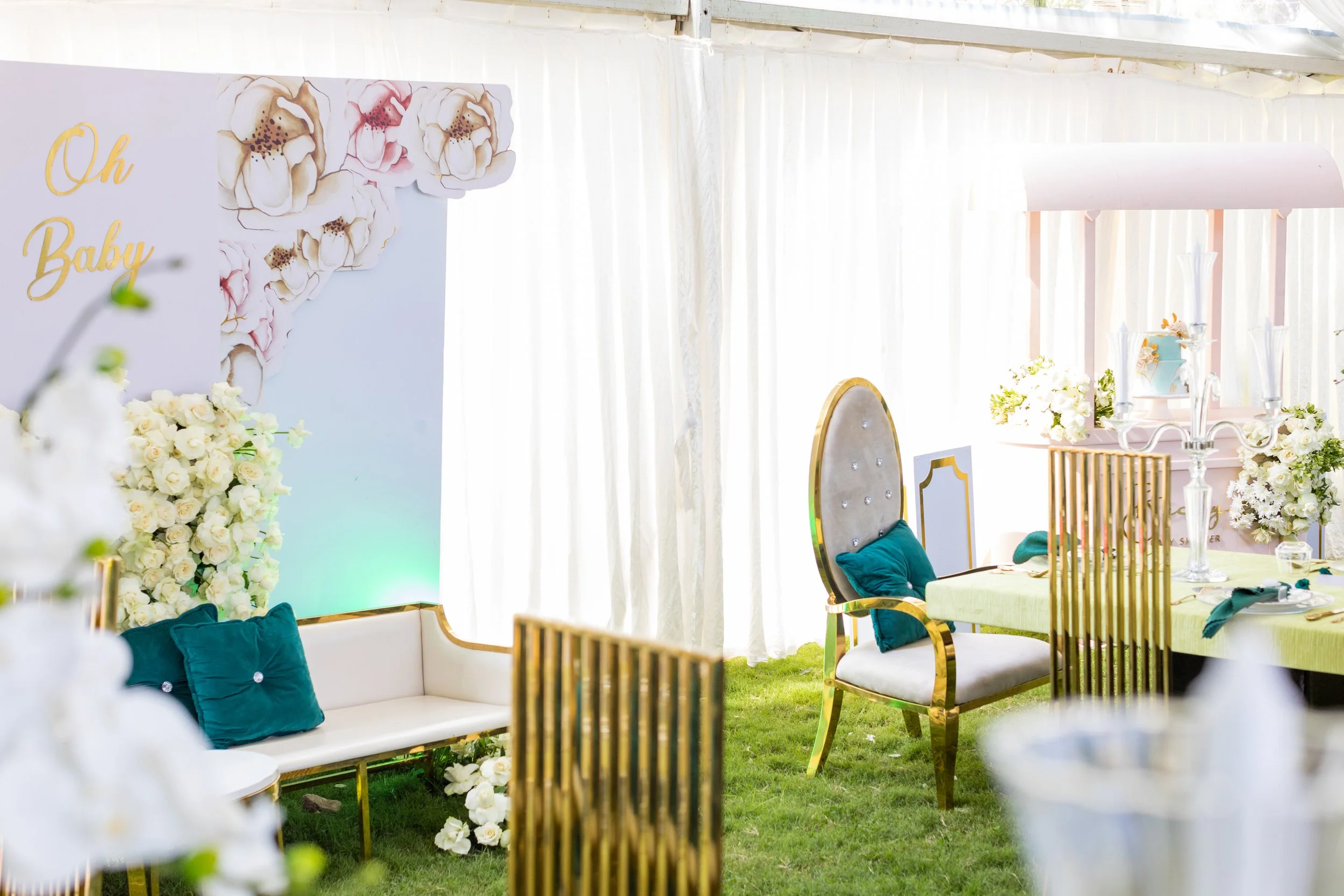 Elegant indoor event space decorated with white and gold accents, floral arrangements, and plush teal pillows on white seating. There is a large floral backdrop with the words 'Oh Baby' written on it.