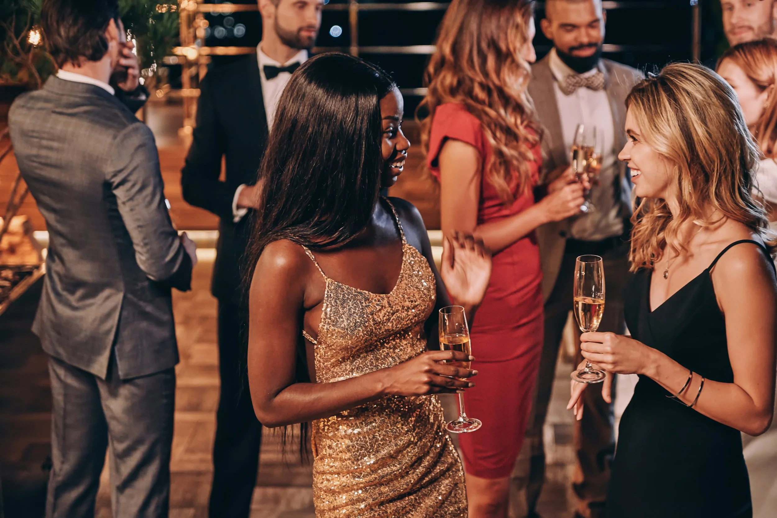 A group of people dressed in formal attire at a social gathering, holding glasses of champagne, engaging in conversation and smiling, in what appears to be a festive event or celebration.