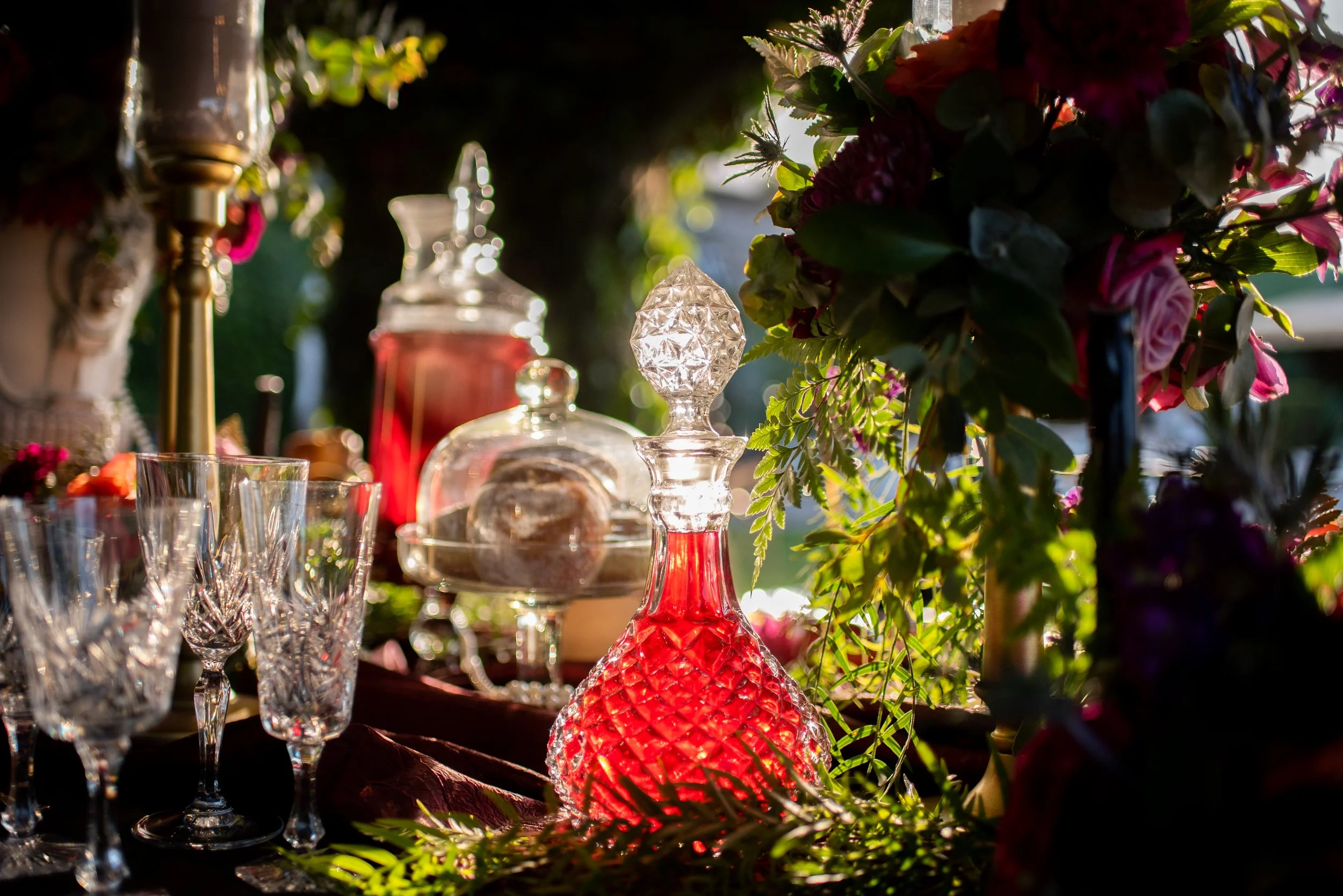A wedding or event decor table with glassware, ornate glass bottles, and a floral arrangement illuminated by sunlight.
