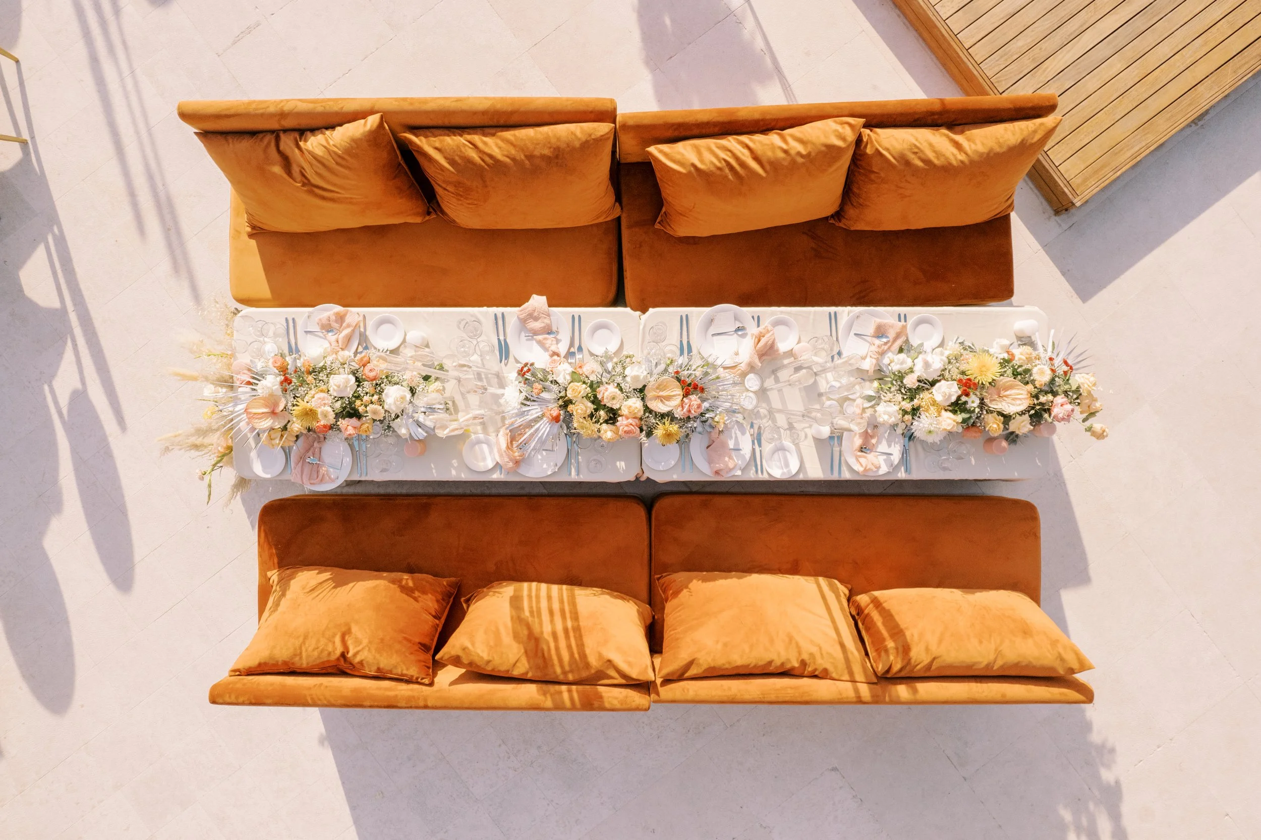 A top-down view of a dining table set for a meal with lush floral centerpieces, surrounded by orange/brown velvet sofas and pillows, on a light-colored tile floor with shadows and a wooden accent wall.