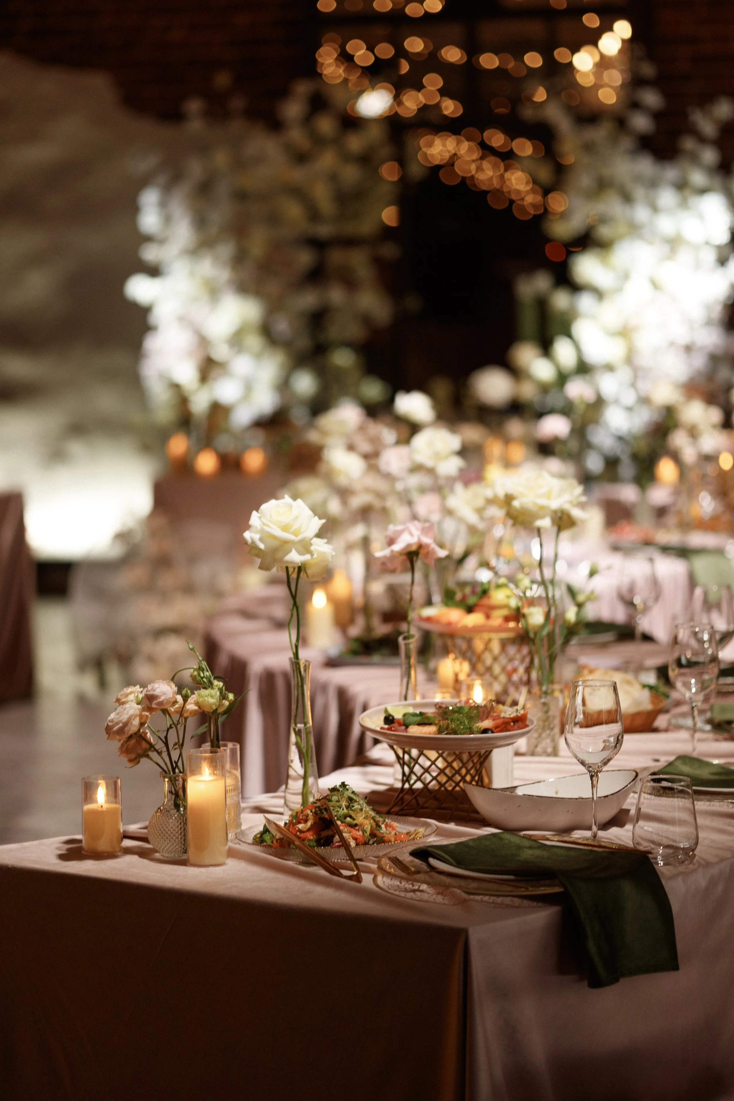 A decorated dining table set for a formal event with candlelit candles, white roses, salad, and glasses, in a warmly lit room with floral and string lights.