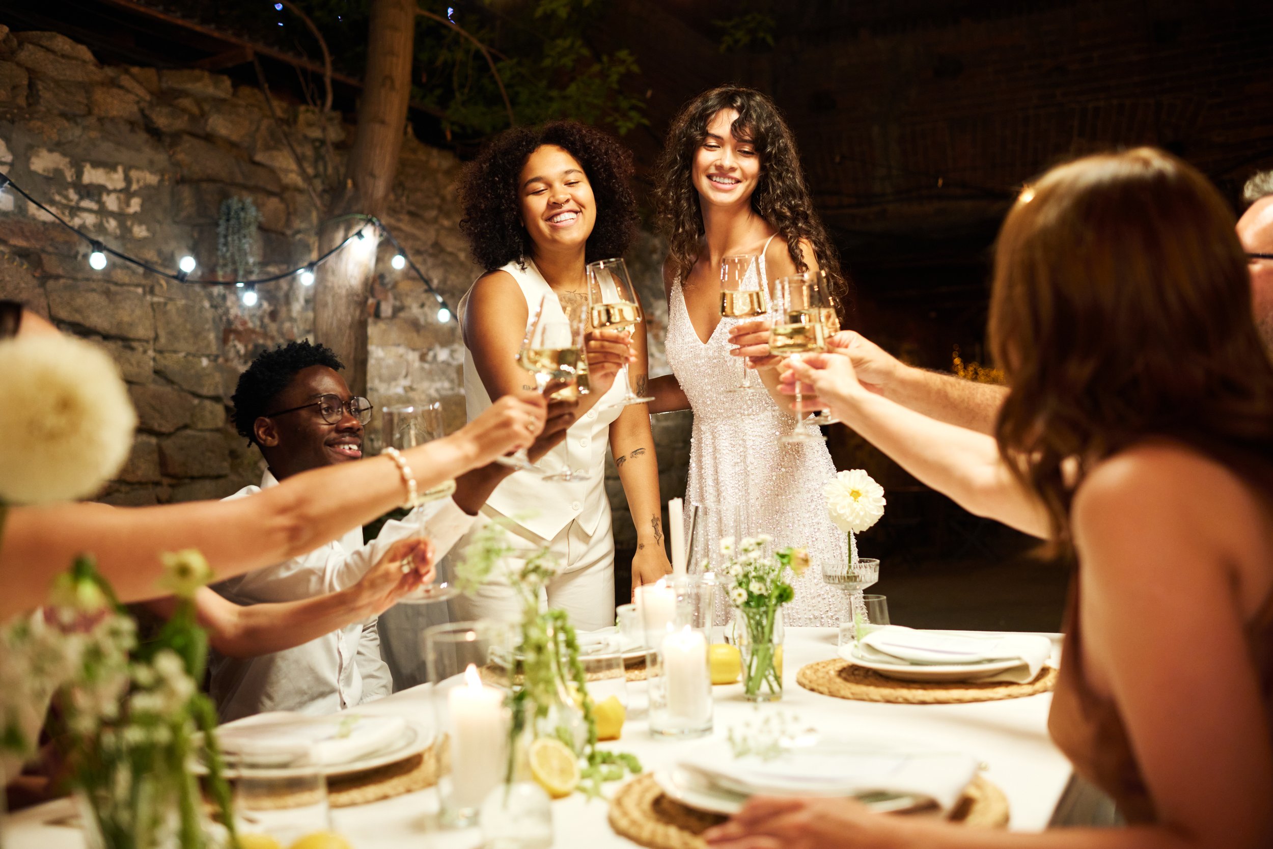 Group of friends celebrating with cheers at a dinner party, with a long table decorated with flowers, candles, and plates, in a rustic setting with string lights.