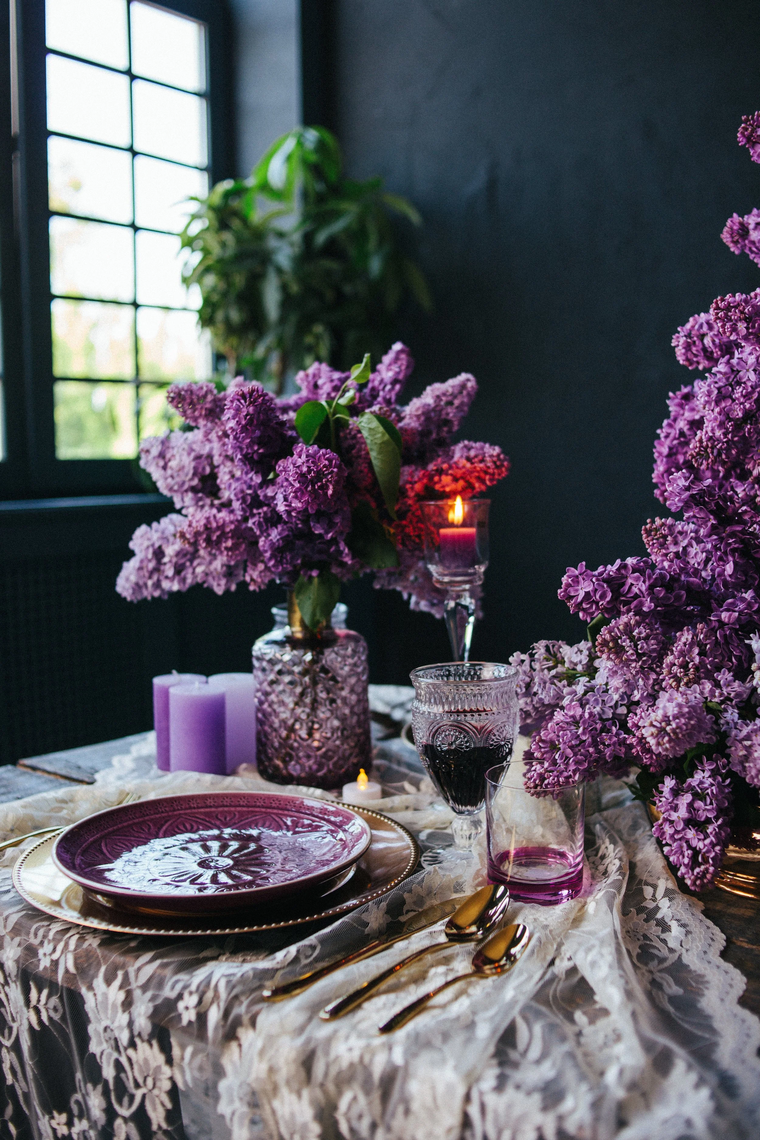 Elegant table setting with purple floral arrangements in glass vases, purple candles, purple glassware, gold flatware, and decorative plates on a lace tablecloth, with a dark wall and window in the background.
