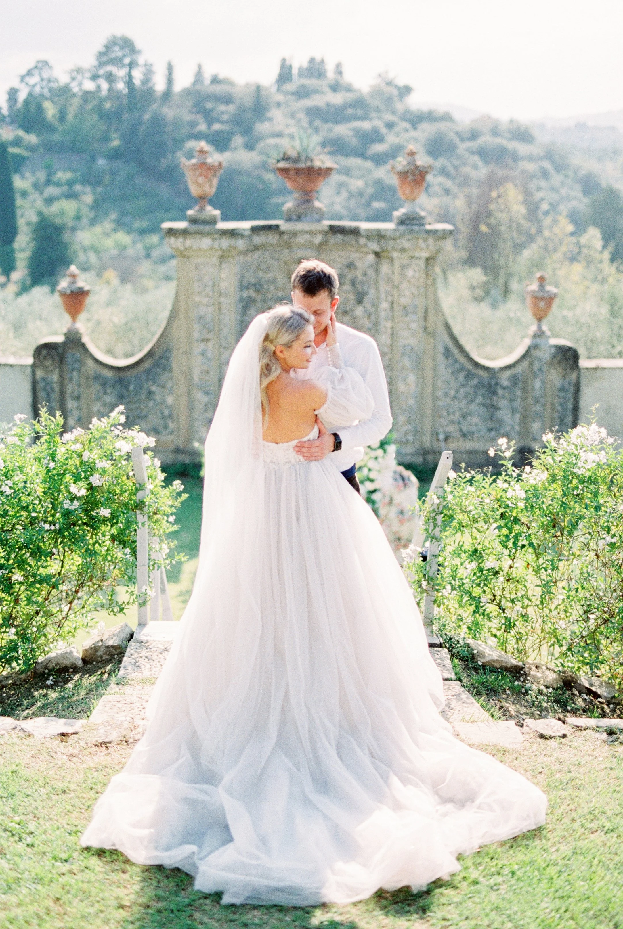 A bride and groom sharing a kiss outdoors during a wedding ceremony, with greenery and a decorative stone wall in the background.