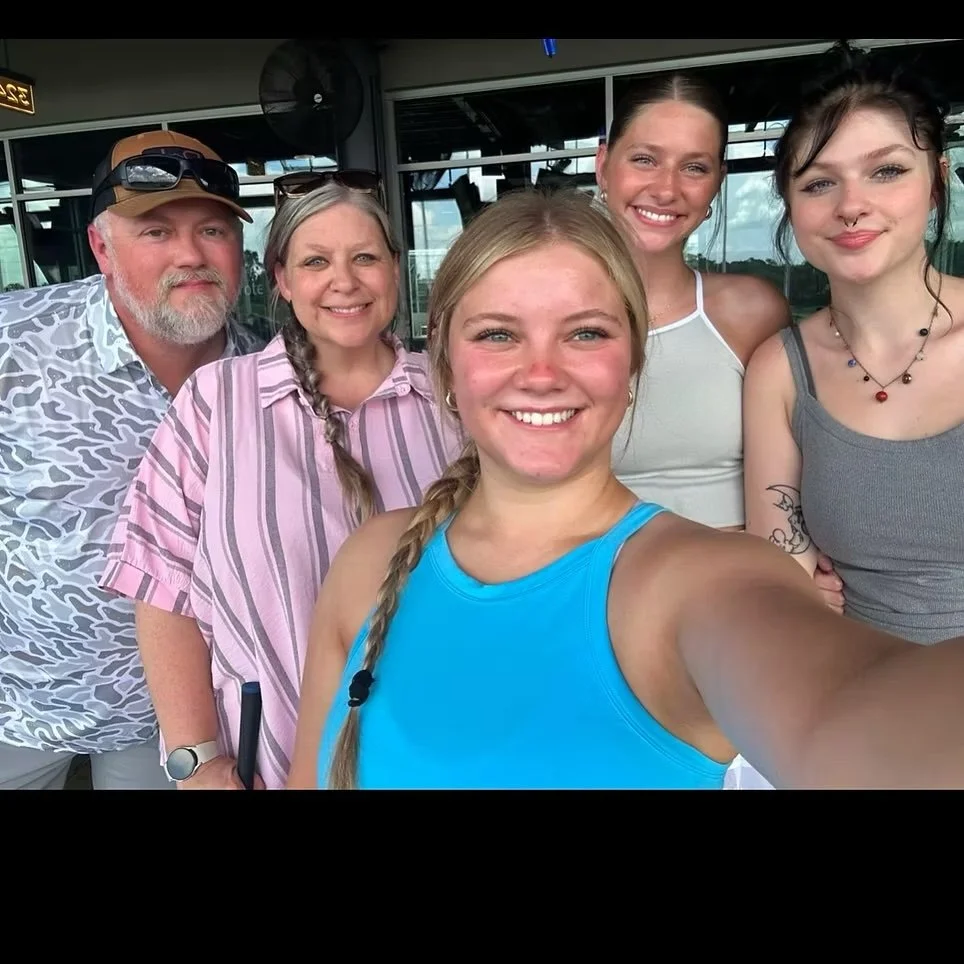 Group of five people smiling for a selfie outdoors, with a man wearing a hat and glasses, woman with braids, and three young women, two with tattoos and necklaces.