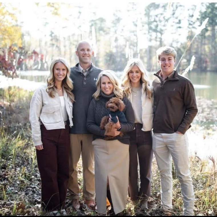 Group of five people and a dog outdoor near a lake or pond with fall foliage in the background