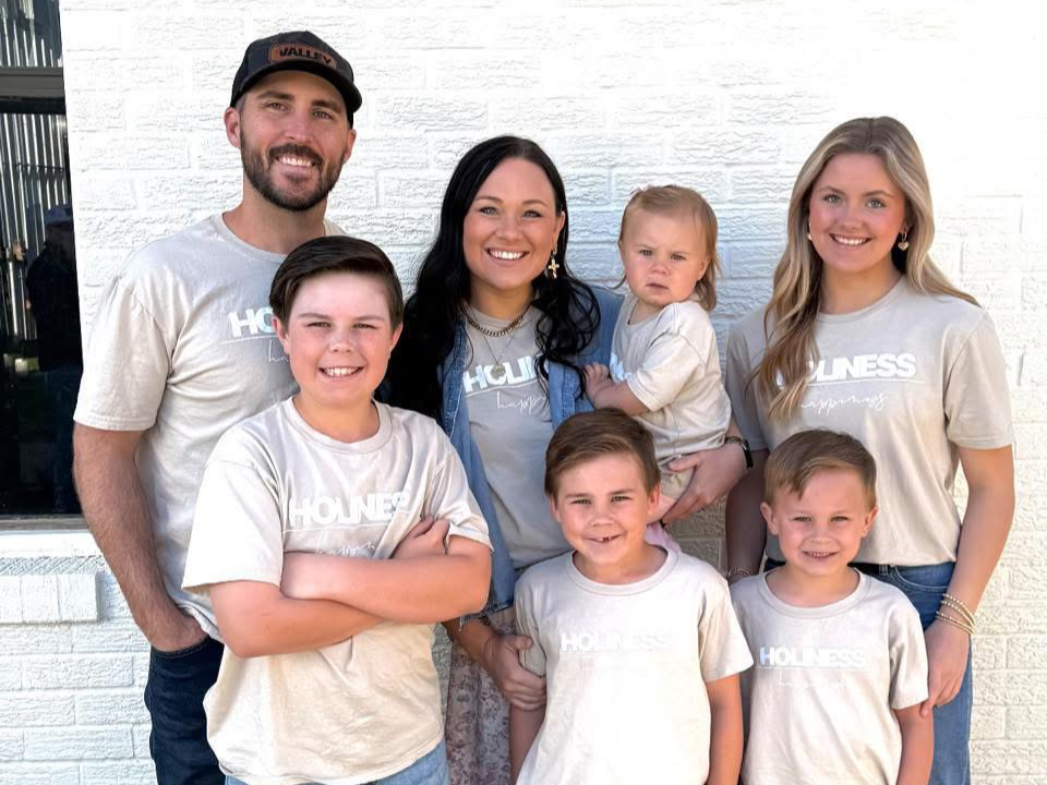 Family of seven standing together outside, smiling, wearing matching beige t-shirts with the word 'HOLINESS' printed on them, against a white brick wall.