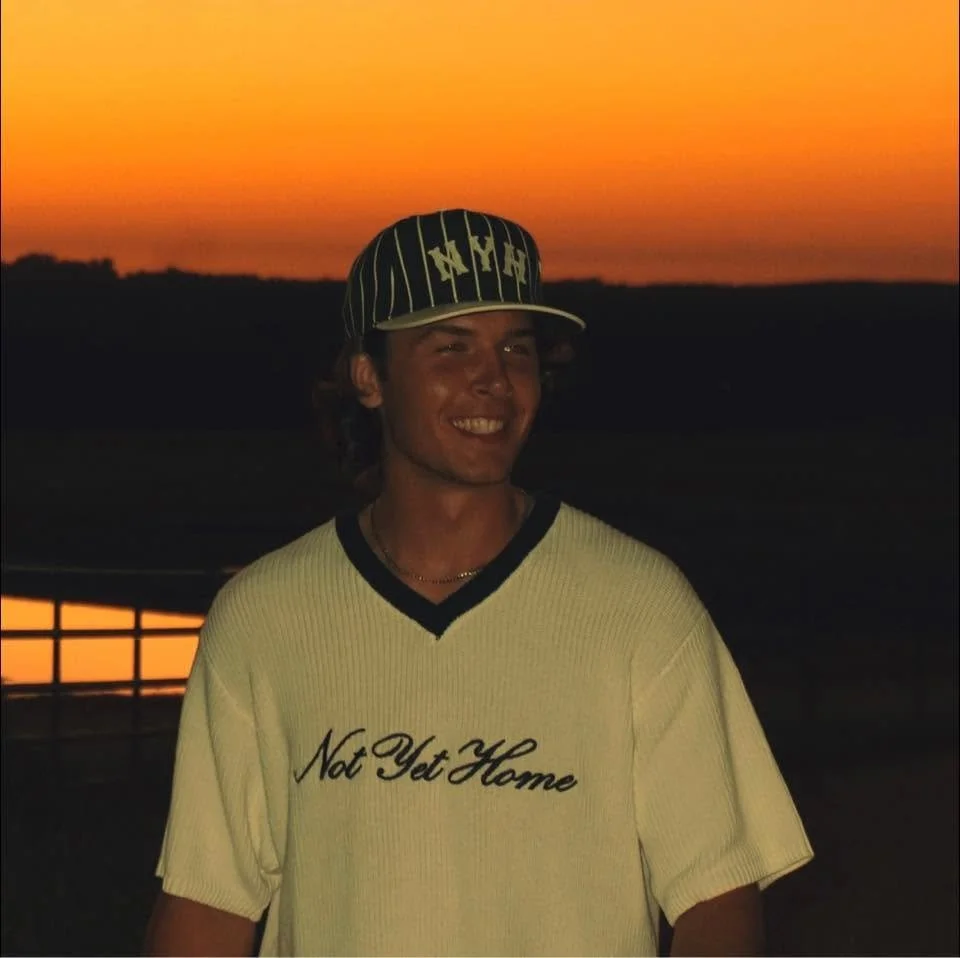 A young man wearing a baseball cap and a cream-colored T-shirt with the words 'Not Yet Home' written on it stands outdoors during sunset, with a dark landscape in the background.