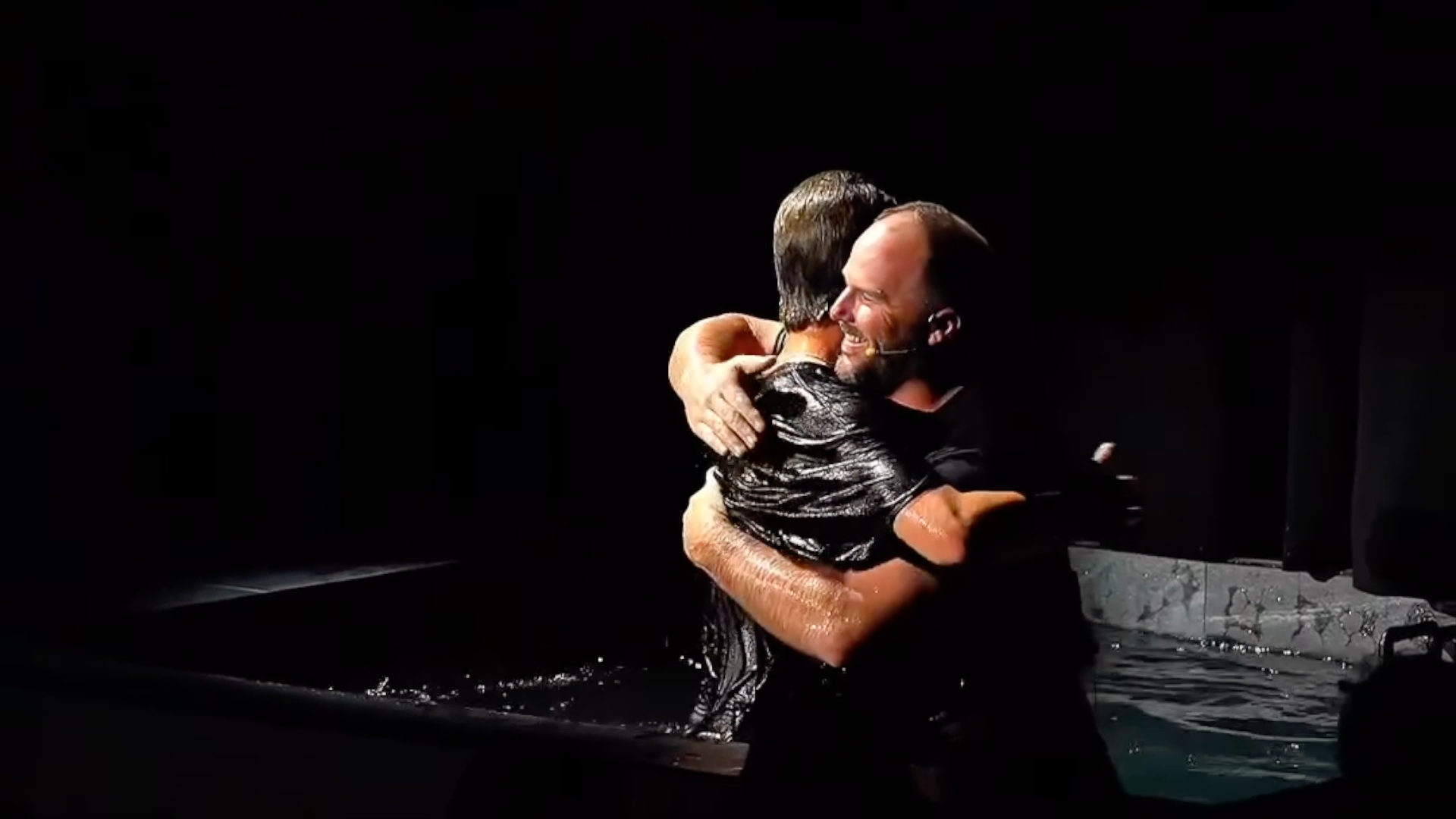 Two men are embracing in a hug in a dark setting, with one man smiling and the other having wet hair and clothing.