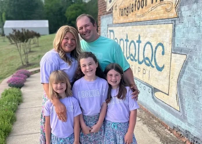 A family of five standing outside next to a wall with a mural. The family includes two adults and three young girls, all smiling and wearing matching purple shirts with a phrase on the front, and patterned skirts or pants.