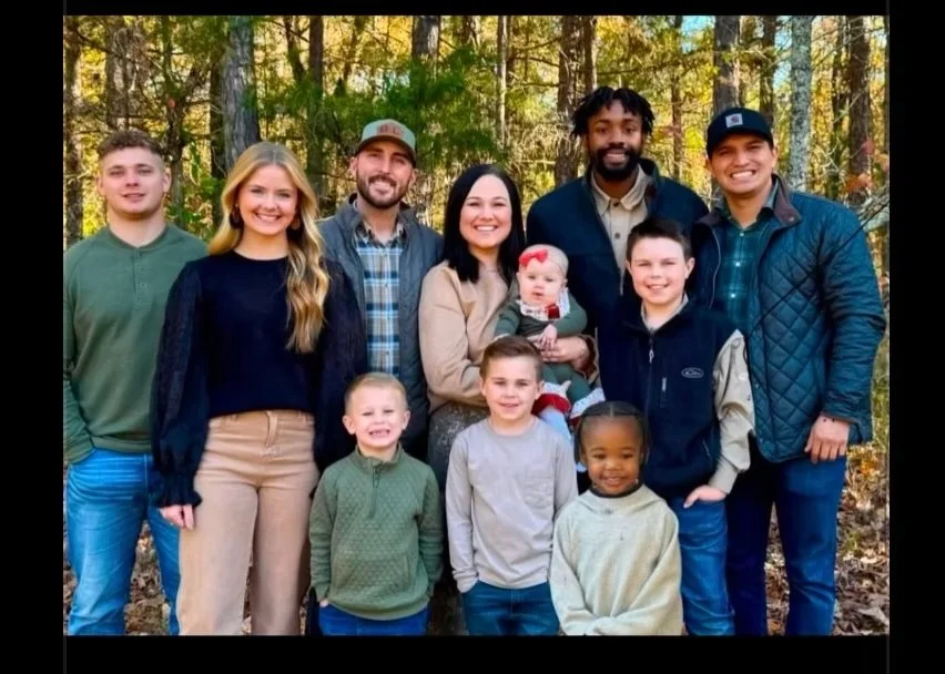 Family photo of nine people, including children and adults, outdoors in a wooded area during daytime.