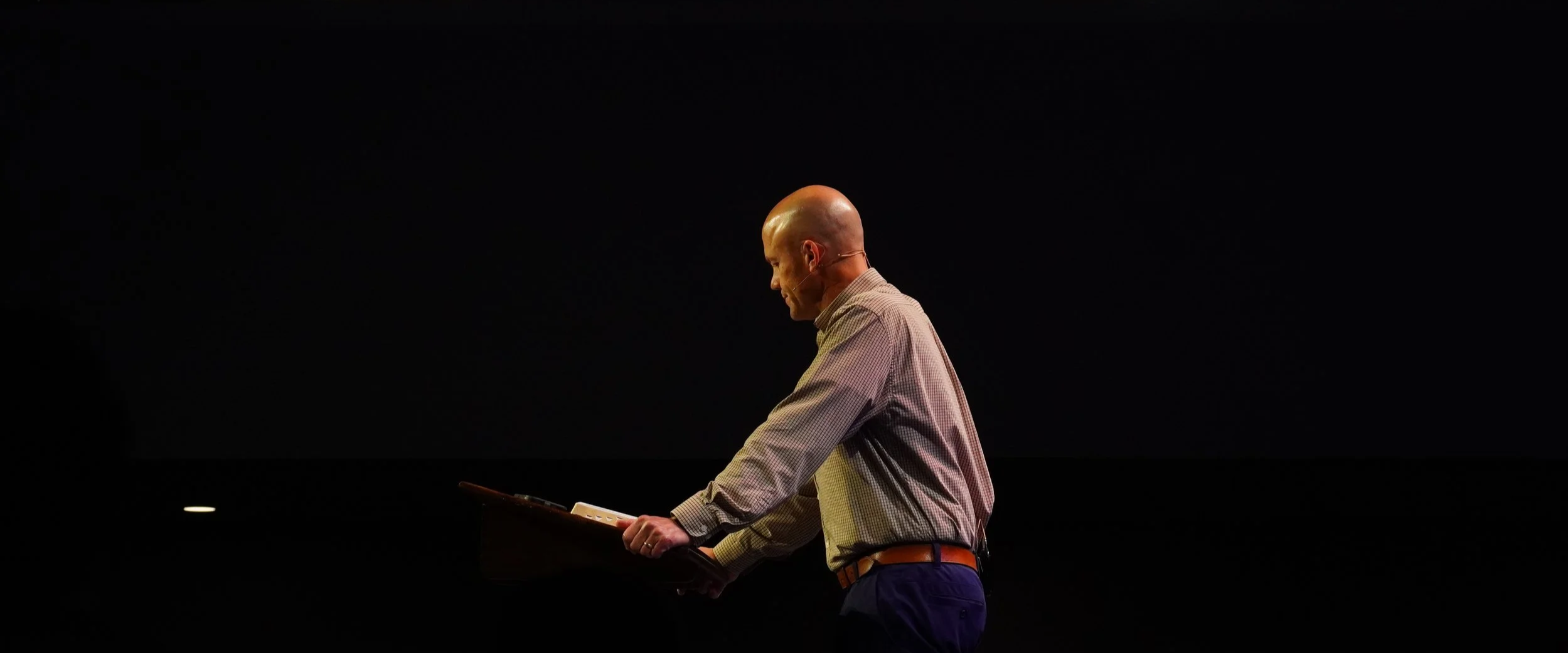 A bald man in a checkered shirt and dark pants stands on a dark stage, looking down at a folder or clipboard in his hands.
