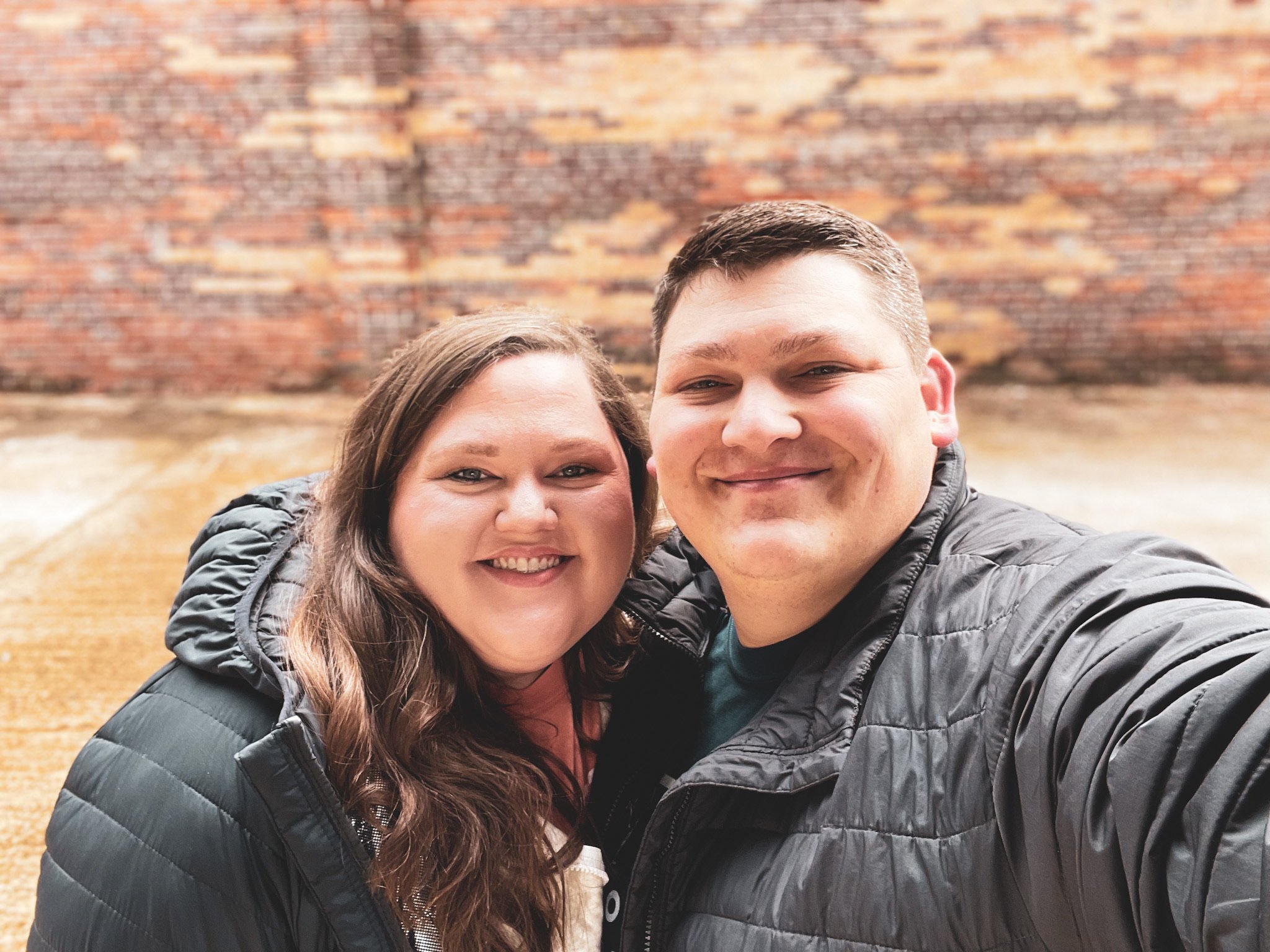 A smiling woman with long brown hair and a man taking a selfie in front of a brick wall with a wooden surface below.