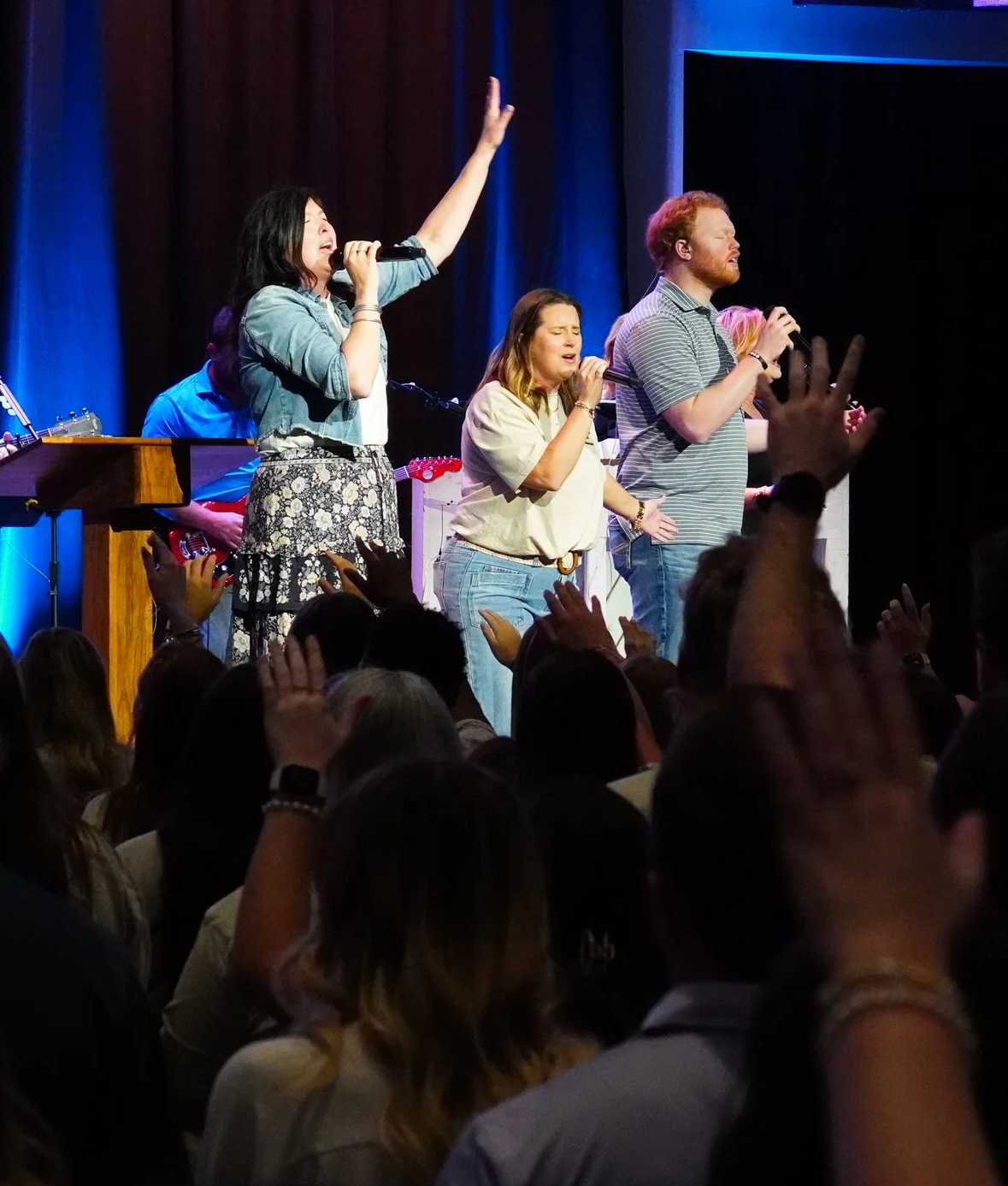 People singing on stage during a concert or religious service with an audience raising hands.