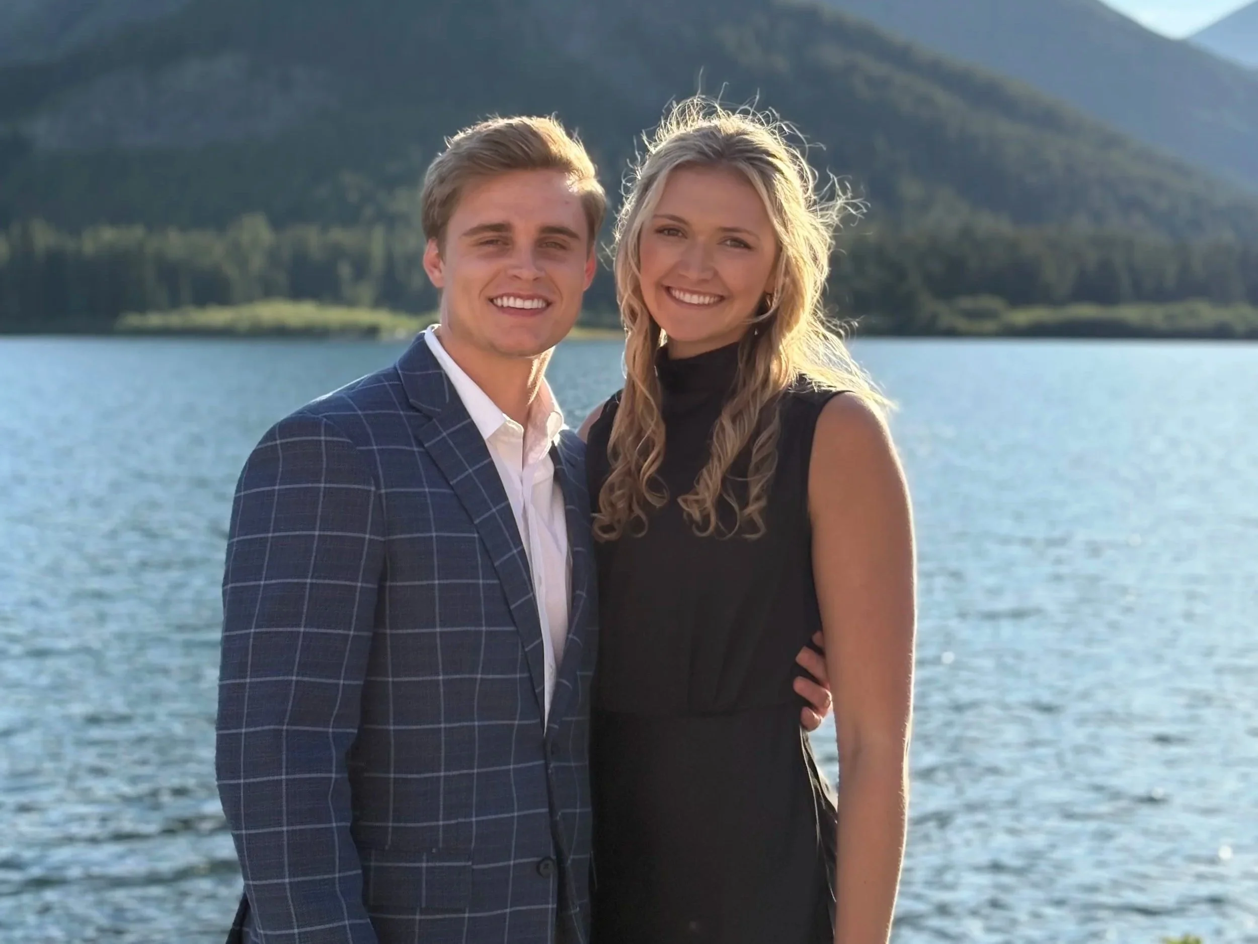 A young couple standing close together outdoors near a body of water with mountains in the background, smiling at the camera.