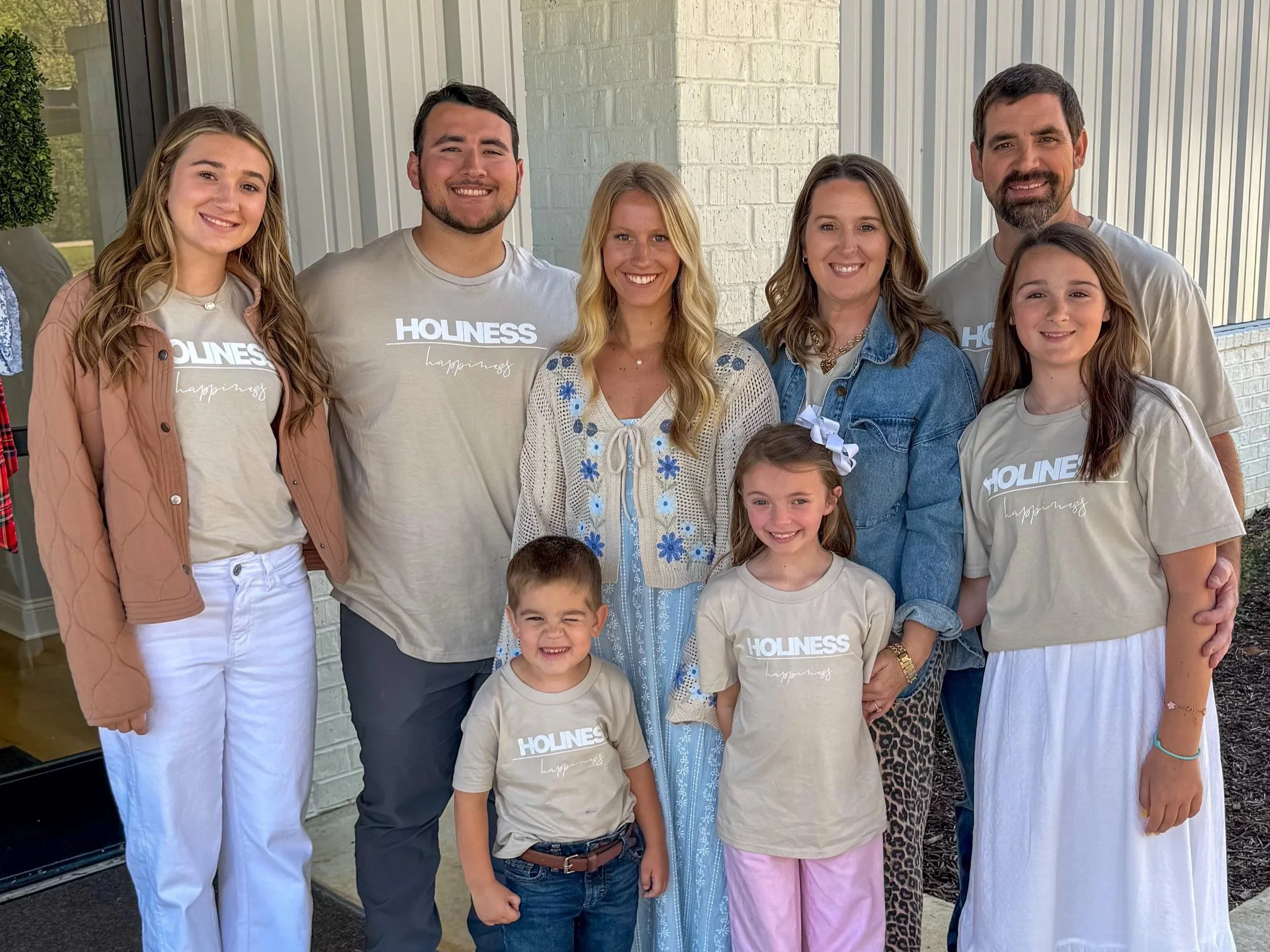 A group of seven smiling people of various ages, standing outside a building with a brick wall and vertical siding, some wearing T-shirts with the words 'HOLINESS' and 'happiness' printed on them.