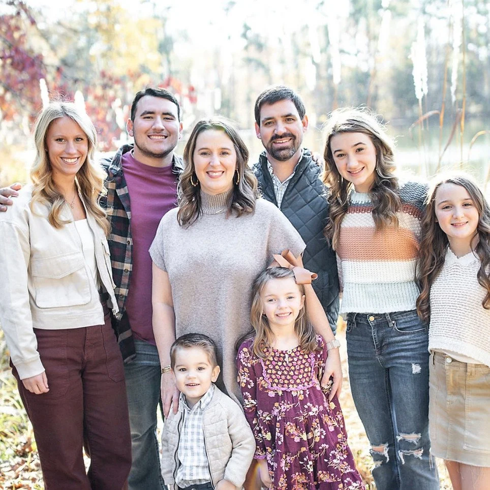 A family of nine people standing outdoors in a park with trees and water in the background, smiling at the camera.