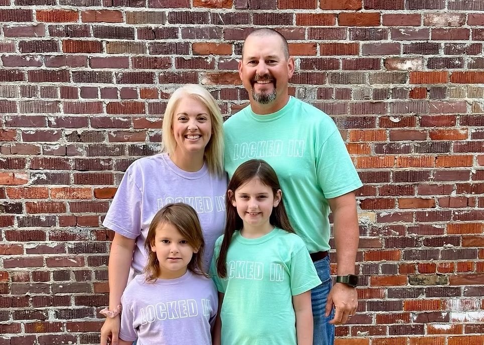 A family of four standing outdoors in front of a brick wall. Two adults, one man and one woman, and two young girls, all smiling and wearing pastel-colored T-shirts.